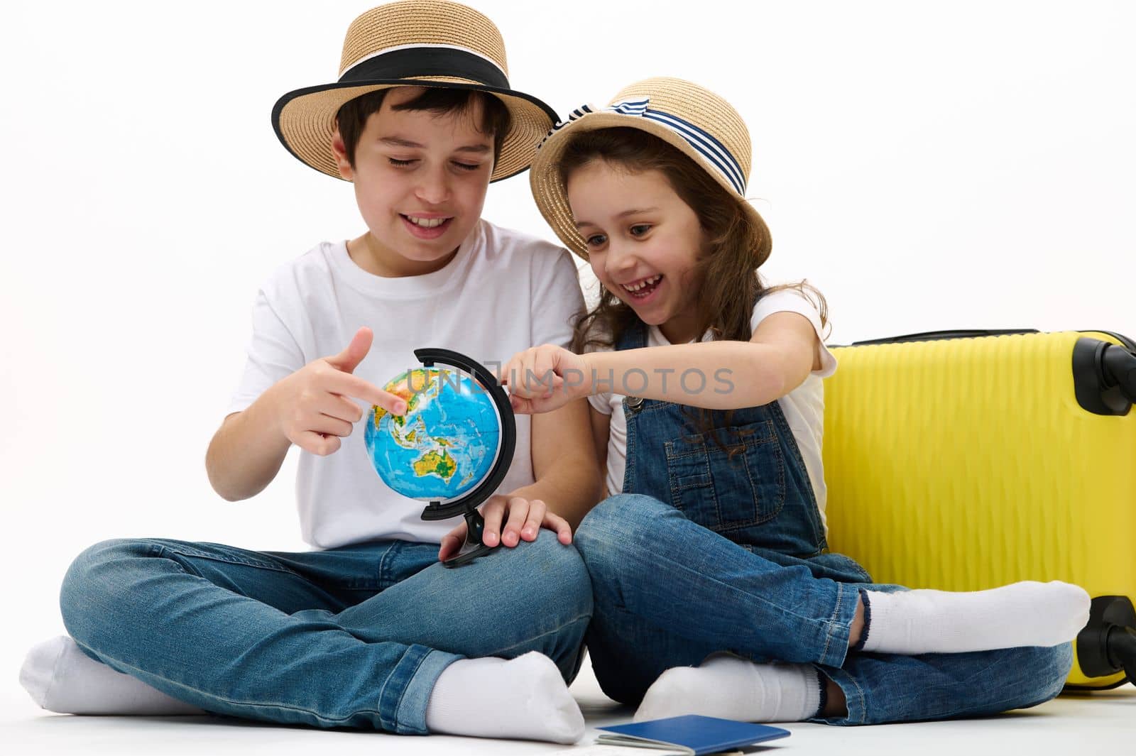 Happy kids, a preteen boy and little girl travelling abroad for summer holidays. Brother and sister pointing at travel destination on the globe map, sitting near yellow suitcase, on white background
