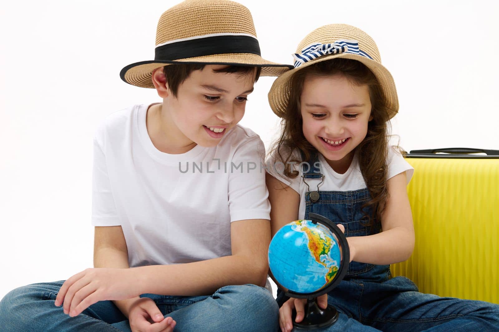 Multi-ethnic travelers kids, boy and girl with suitcase, brother and sister going for summer vacations, pointing fingers at place on globe with Earth map, choosing new travel destination for holidays