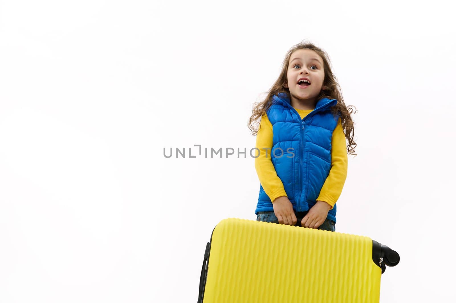 Overjoyed Caucasian little traveler girl wearing yellow pullover and blue down jacket, carrying a heavy yellow suitcase, posing with open mouth and surprised look over white background with copy space