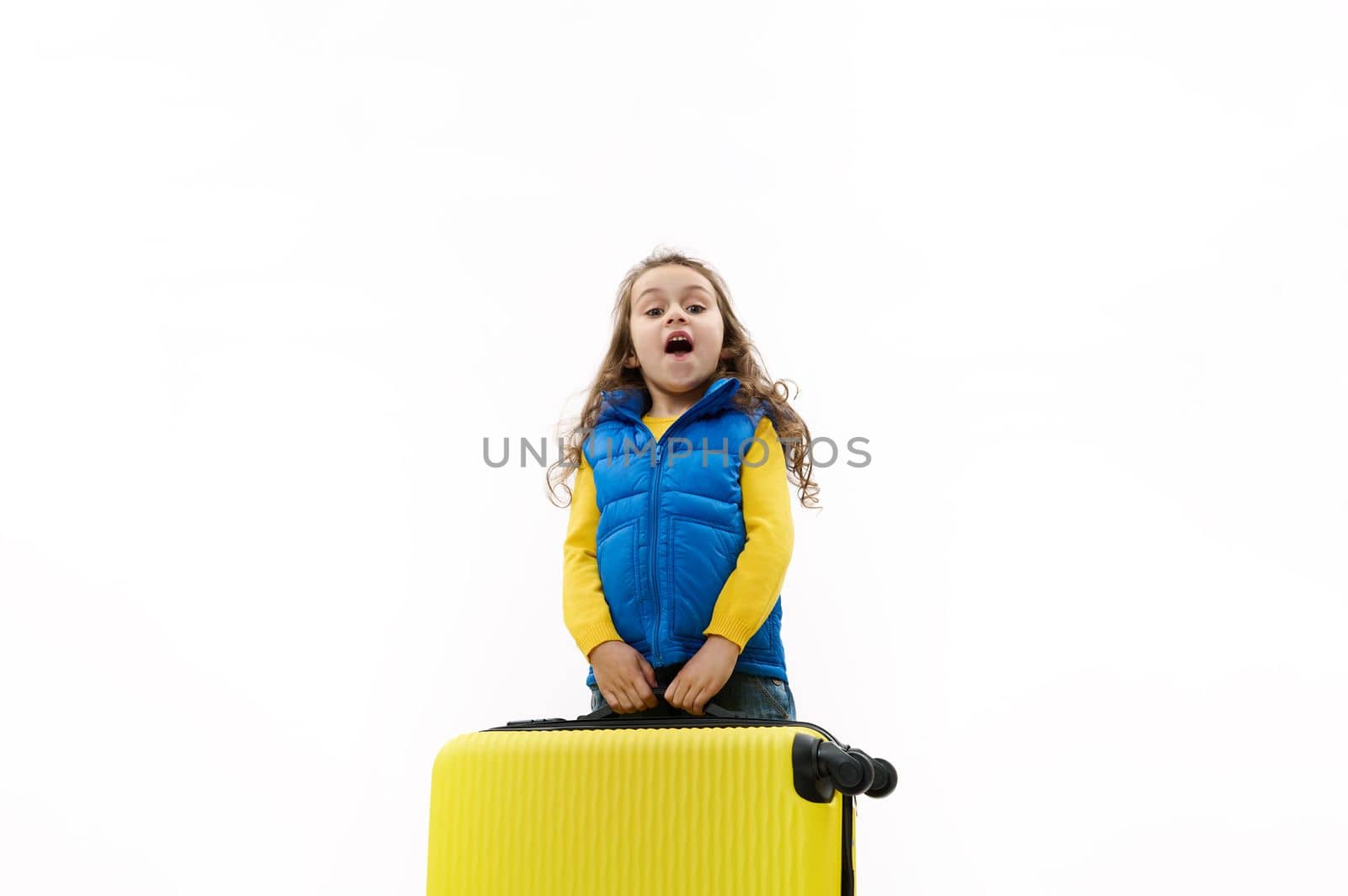 Caucasian beautiful little kid girl in blue jacket, going for weekend getaway, carrying a heavy yellow suitcase, isolated on white background. Travel. Trip. Journey. Air Flight. Copy advertising space