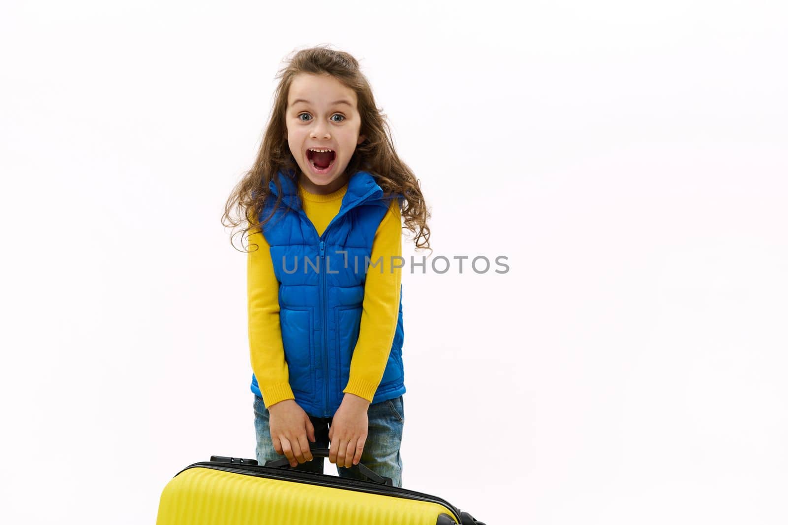 Mischievous little traveler kid girl 5-6 years old, holding yellow suitcase, isolated on white background. Passenger traveling abroad on weekends getaway. Air flight journey concept. Copy ad space
