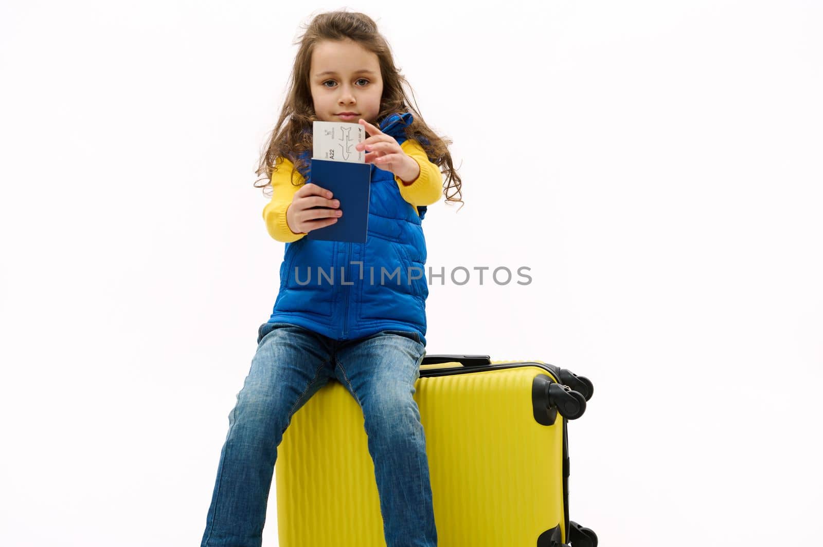 Isolated portrait on white background of a Caucasian charming kid girl in blue jacket, holding out an air ticket and boarding pass at camera, sitting on yellow travel bag, going for weekend getaway.