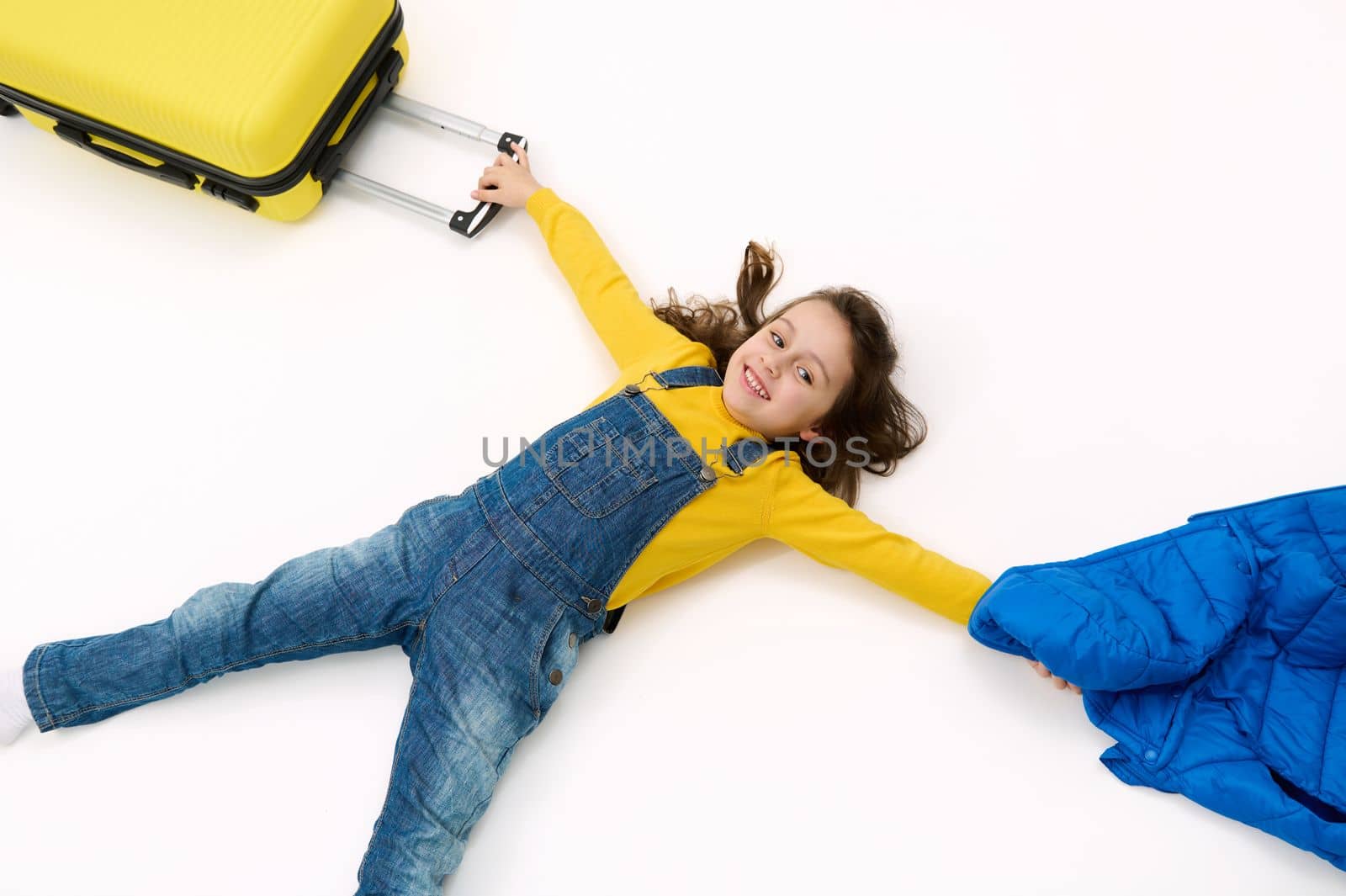 Top view little child girl tourist in blue denim overalls, with yellow sweater and suitcase, lying on white background, by artgf