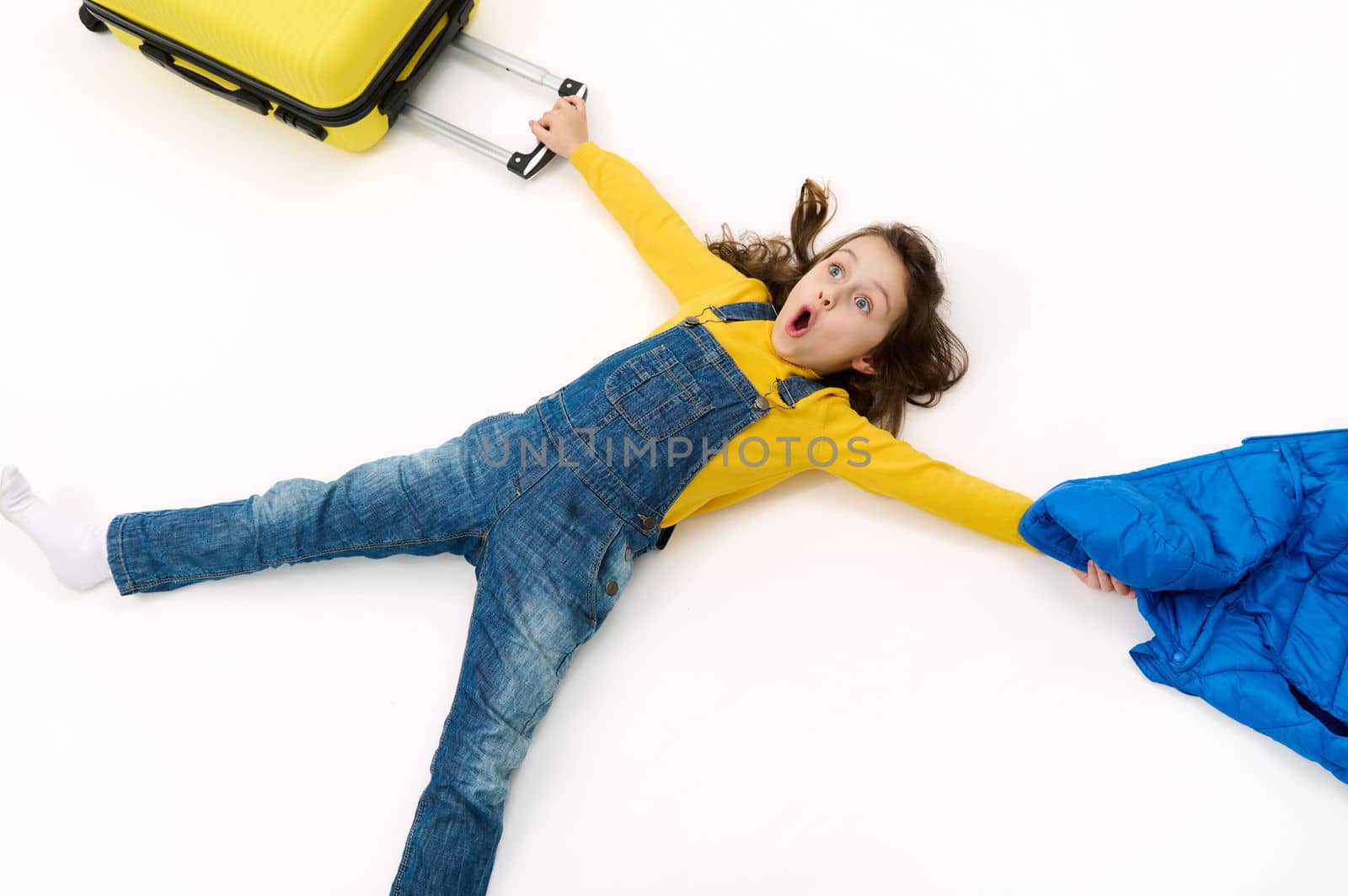 Surprised little girl in yellow sweater and blue denim overalls holding her coat and suitcase in outstretched hands, expressing wow emotion, amazement and astonishment, lying down on white background