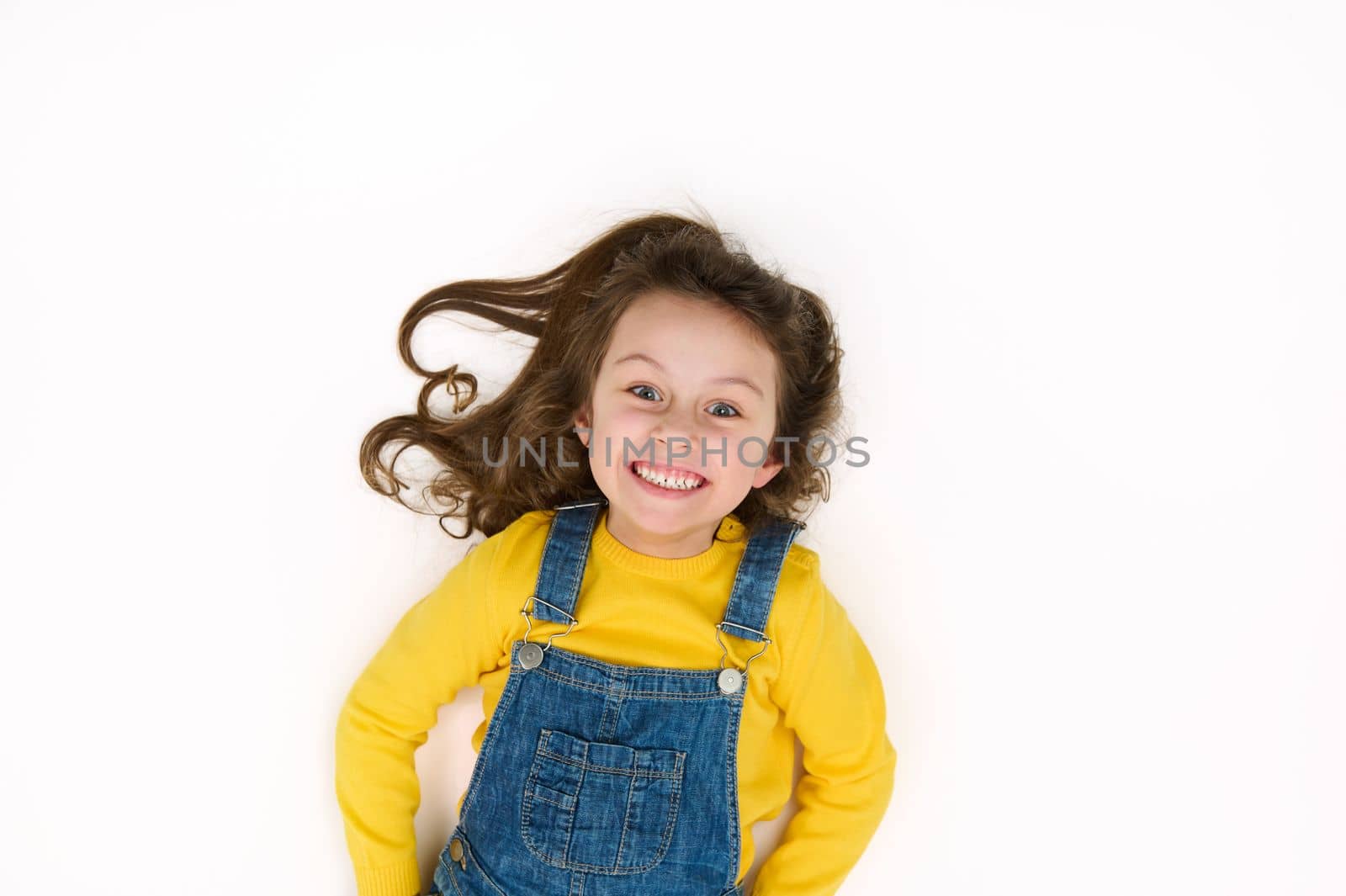 Charming European 5-6 years little child girl, in yellow sweater and blue denim overalls, smiles a cheerful toothy smile, looking at camera, isolated over white background with copy advertising space