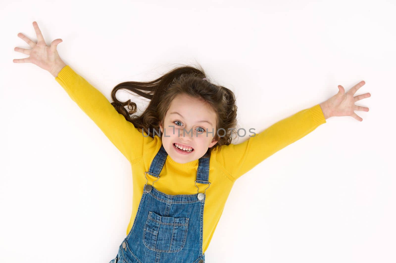 Overhead view of an adorable mischievous Caucasian baby girl with outstretched arms, making faces, rejoicing, smiling, looking at camera while lying on white background with copy advertising text