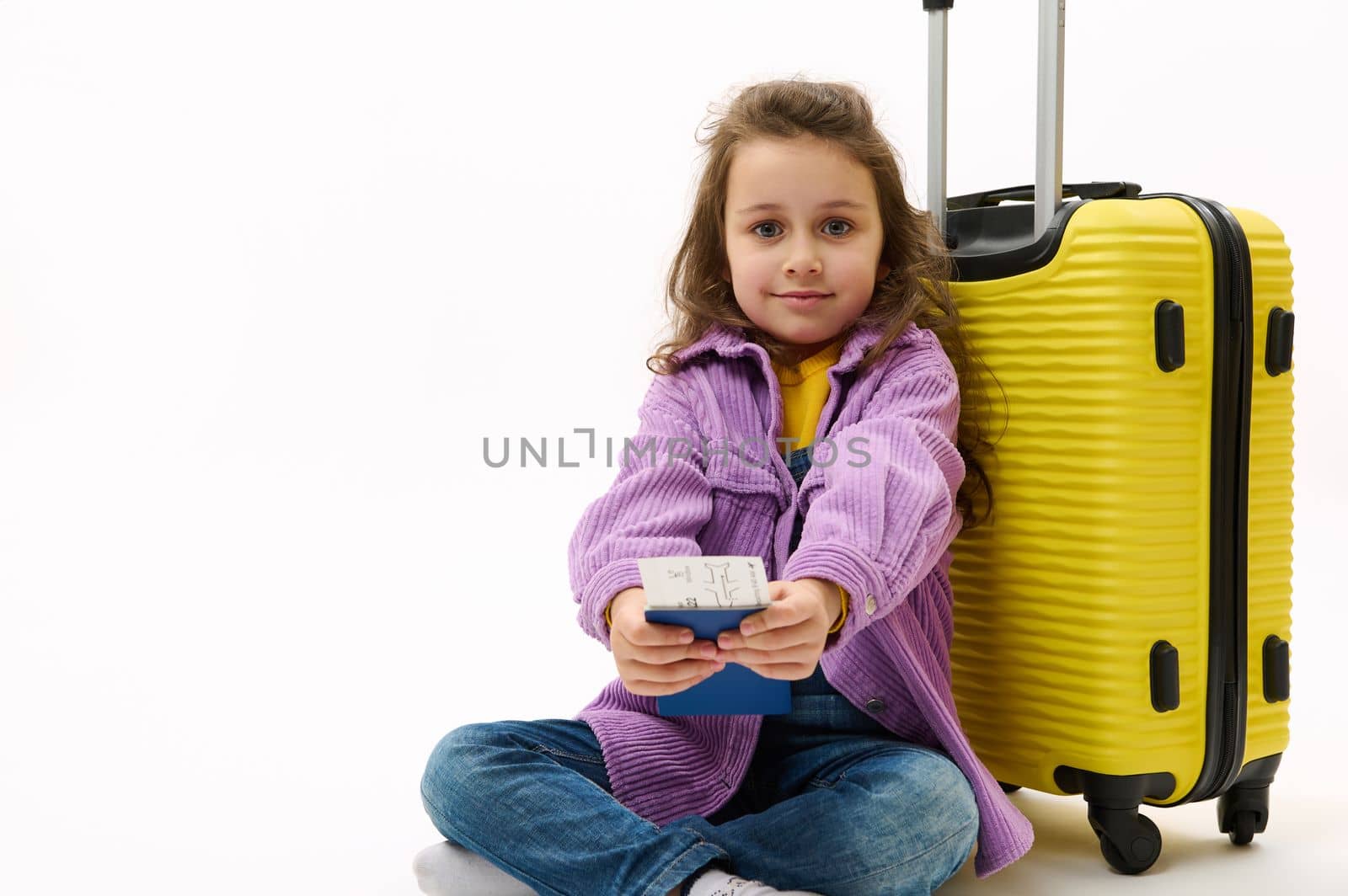 Lovely little traveler girl wearing purple shirt and denim overalls, holding plane ticket sitting near a yellow valise on white background with free advertising space. Kid travels abroad for vacations