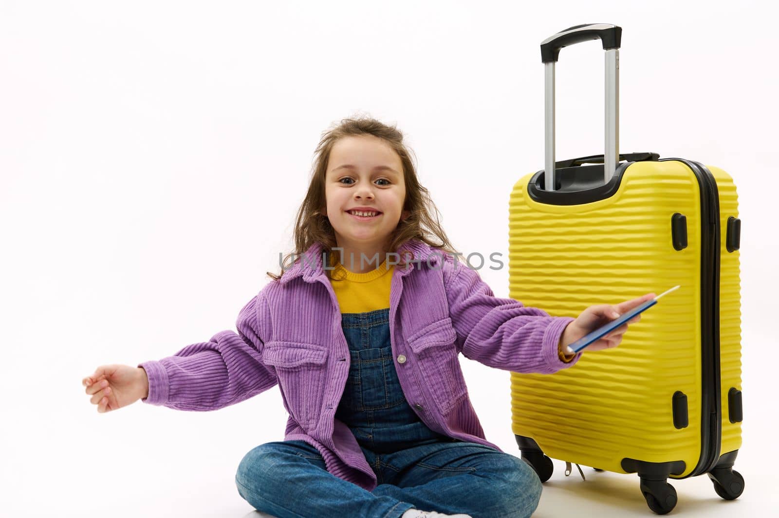 Cheerful Caucasian traveler child girl in purple shirt and blue denim overalls, smiling a beautiful toothy smile looking at camera, posing with boarding pass and yellow suitcase on white background