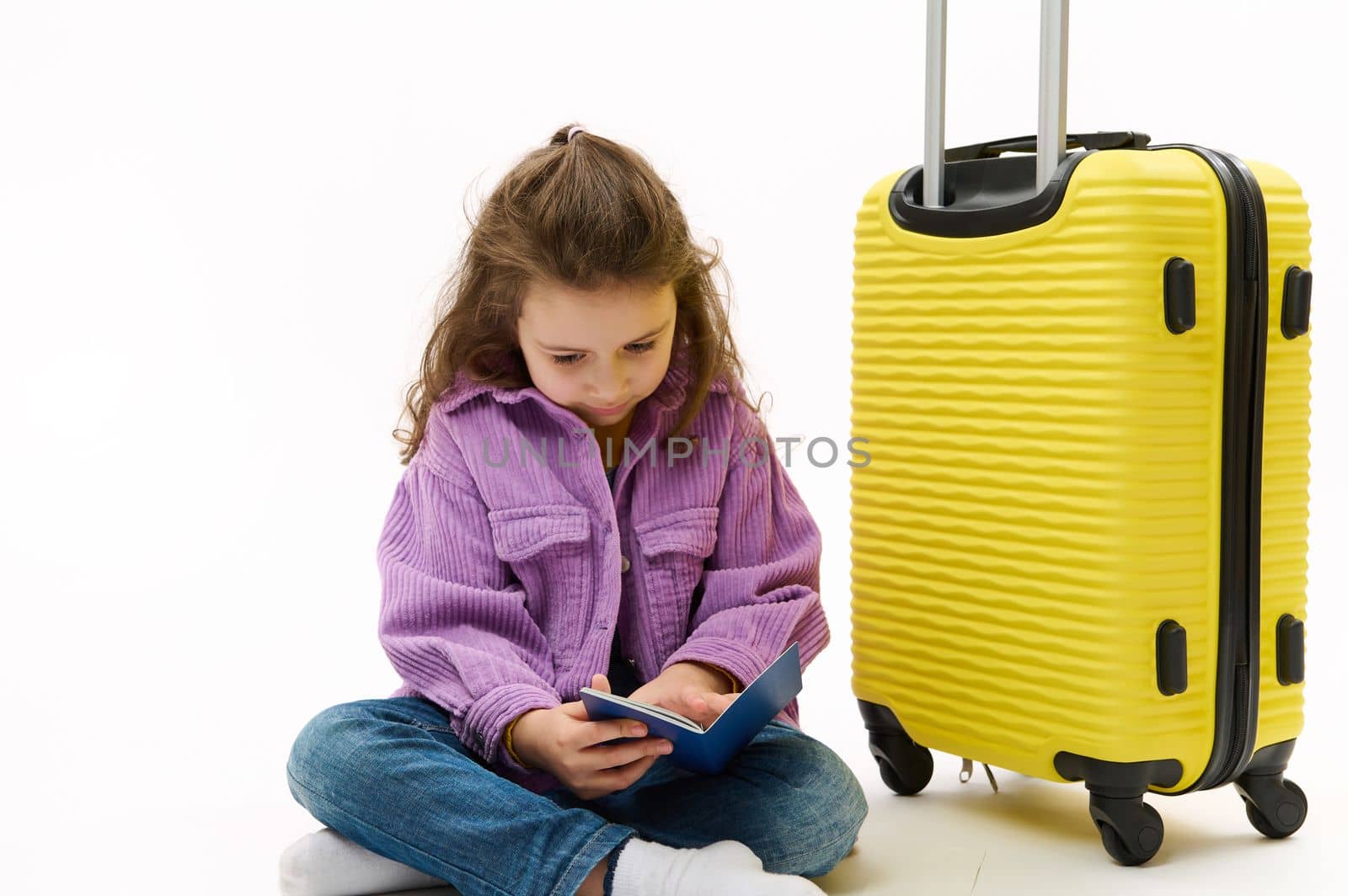 Adorable little child, traveler baby girl wearing blue jeans and purple shirt, sitting near a yellow suitcase with boarding pass in her hands, going for weekend getaway, isolated over white background