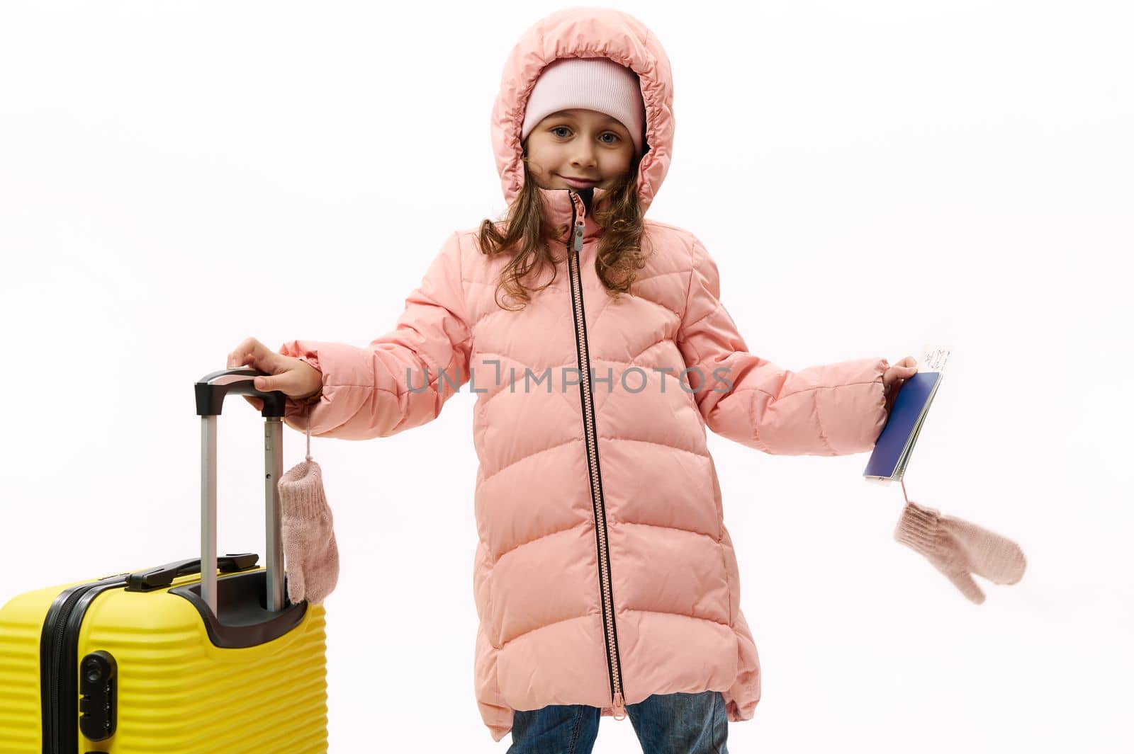 Charming little traveler girl wearing pink warm down coat, standing near a yellow suitcase with boarding pass in her hands, going for weekend getaway, isolated over white background. Winter holidays