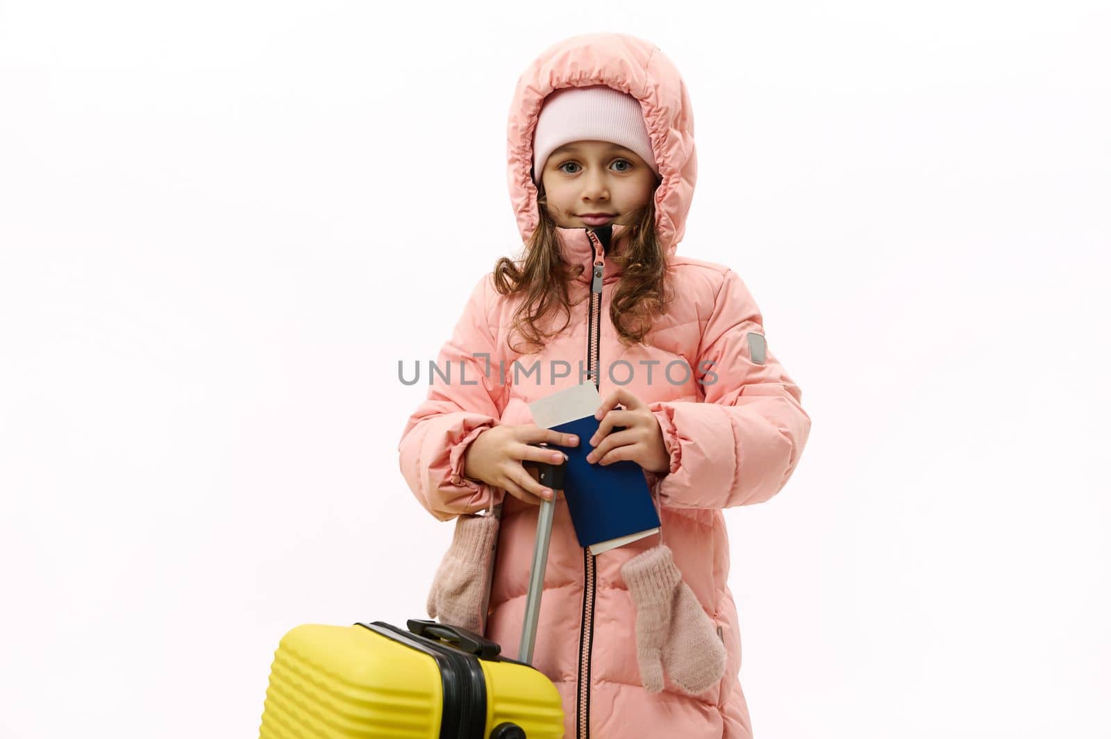 Beautiful Caucasian little girl in pink down coat, going for vacations, looking at camera, posing with suitcase and boarding pass on white background with free advertising space. Travel Trip Journey