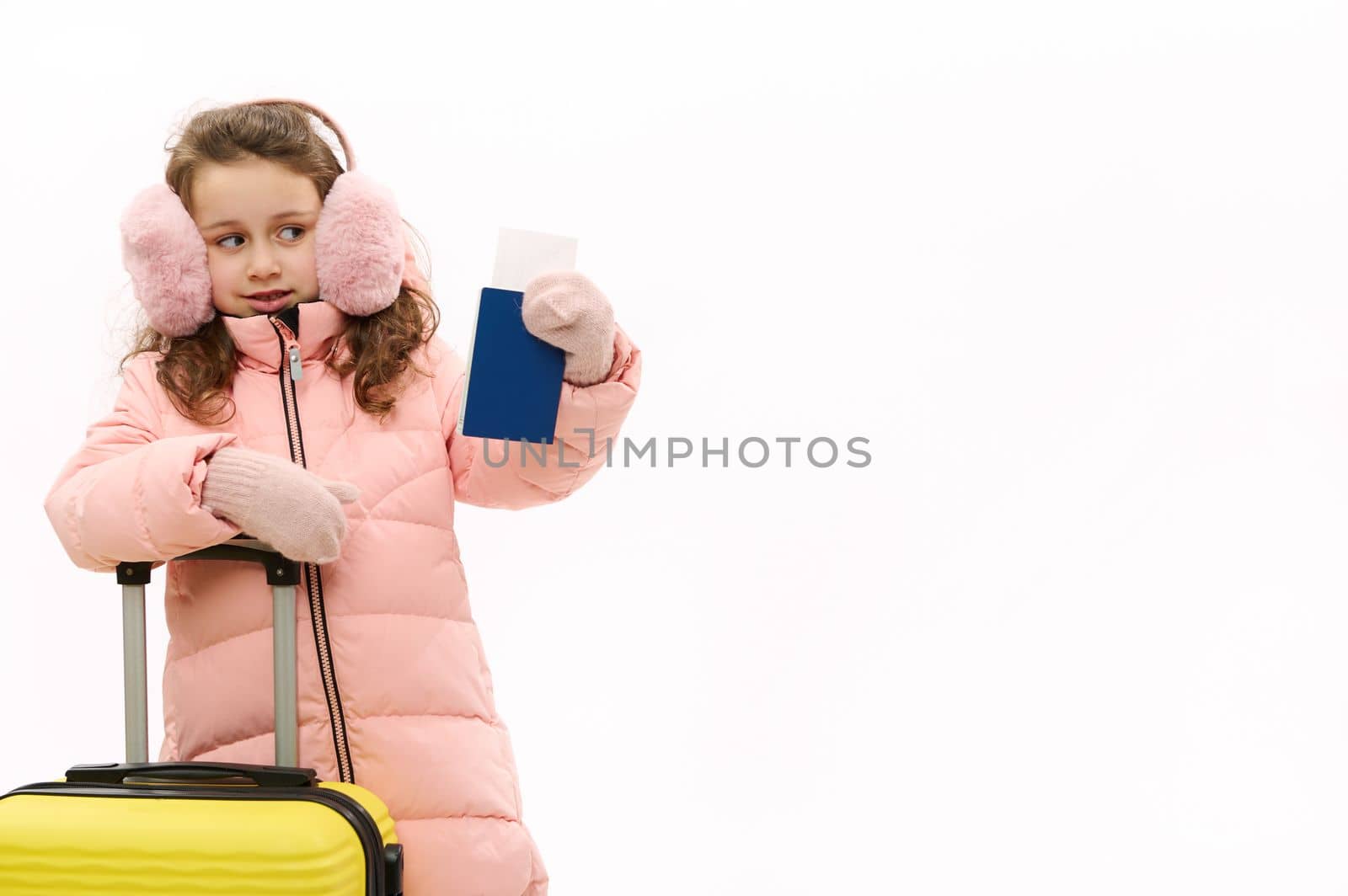 Adorable little traveler girl in warm winter clothes and fluffy headphones, going for weekend getaway, posing with boarding pass and suitcase, looking at copy space on white background. Journey Travel