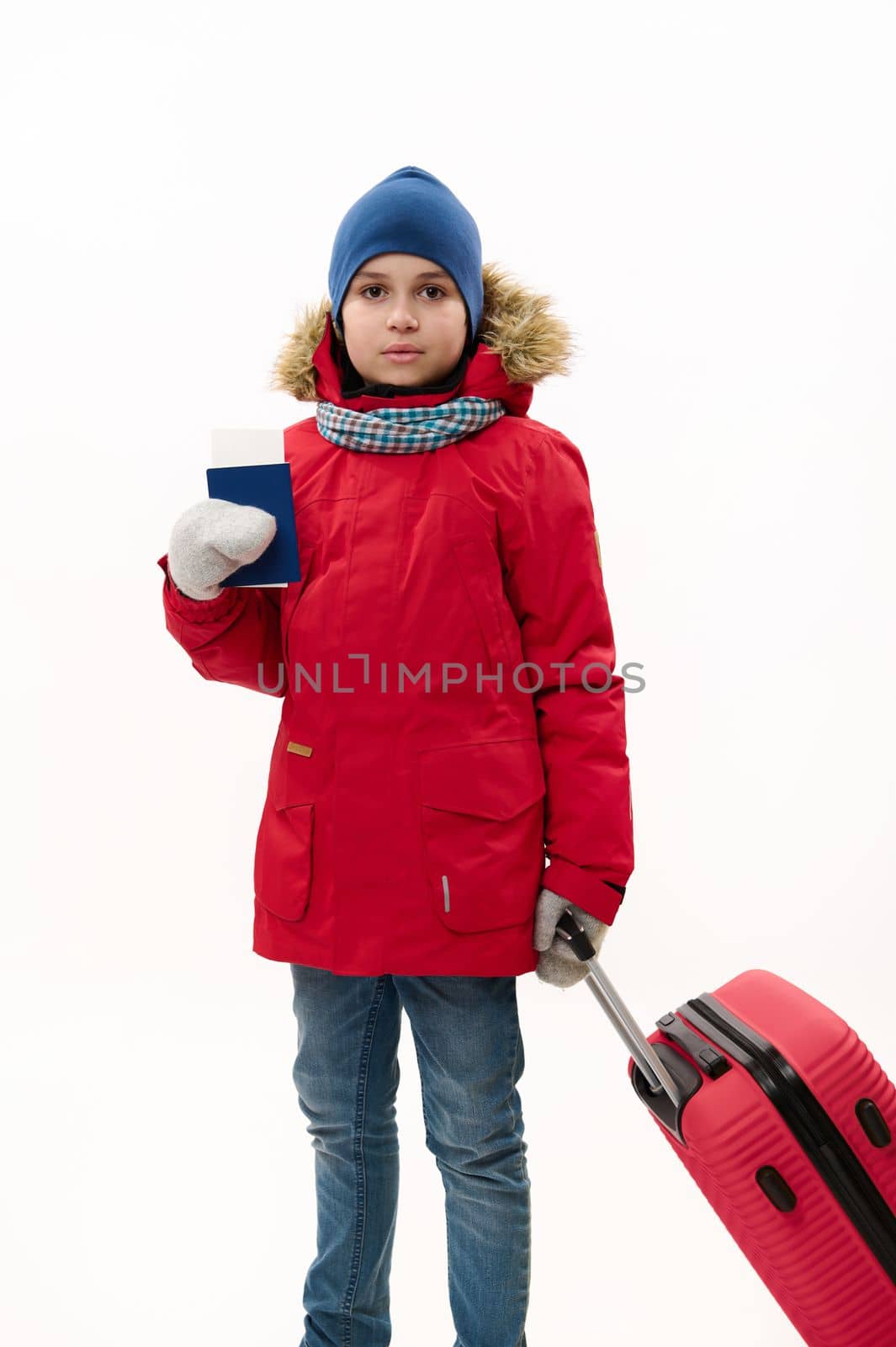 Full length portrait of teenage traveler boy wearing down jacket and blue jeans, going for winter vacations, walking with boarding pass and red suitcase over white background. Journey. Travel concept