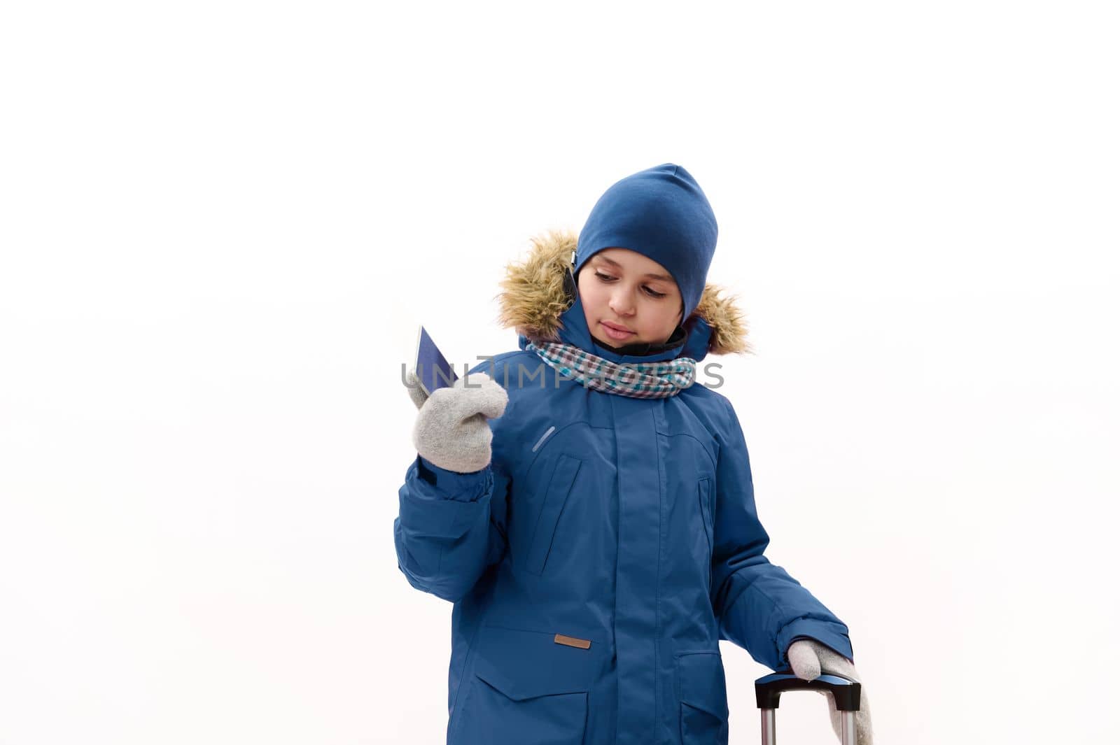 Adorable preteen child, traveler boy in blue parka and wool mittens, going for winter holidays, holding a boarding pass and travel bag, isolated over white background. Rest getaway, journey concept