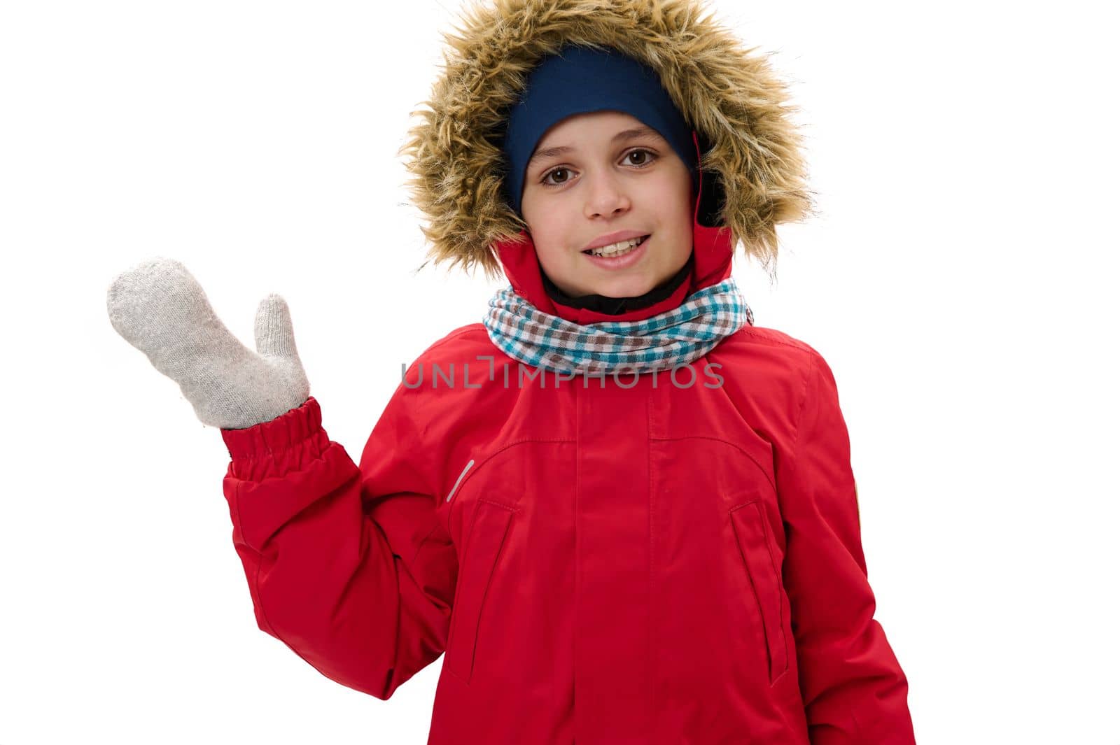 Adorable Caucasian teen boy in warm winter clothes, waves hello with hand, smiles a cheerful toothy smile, looking at camera, isolated on white background with copy space for your advertising text