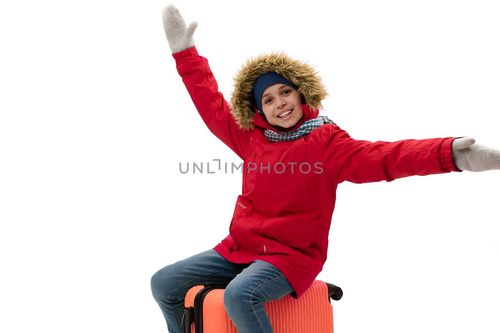 Caucasian smiling teenage traveler boy, wearing warm red down jacket, wool mittens and blue jeans, sitting on orange suitcase, travels abroad, isolated over white. Kids travelling on weekends getaway