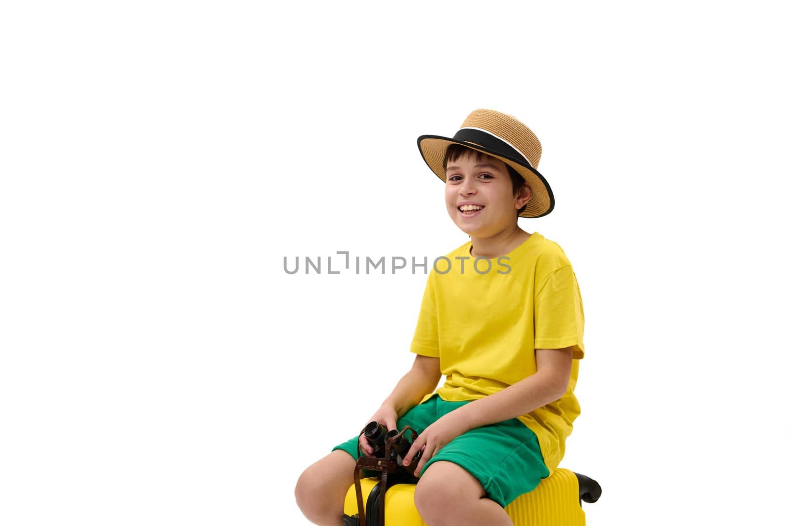 Charming teenage boy wearing straw hat, yellow t-shirt and green shorts, going for summer holidays, looking at camera, sitting on suitcase with binoculars in his hands, isolated over white background