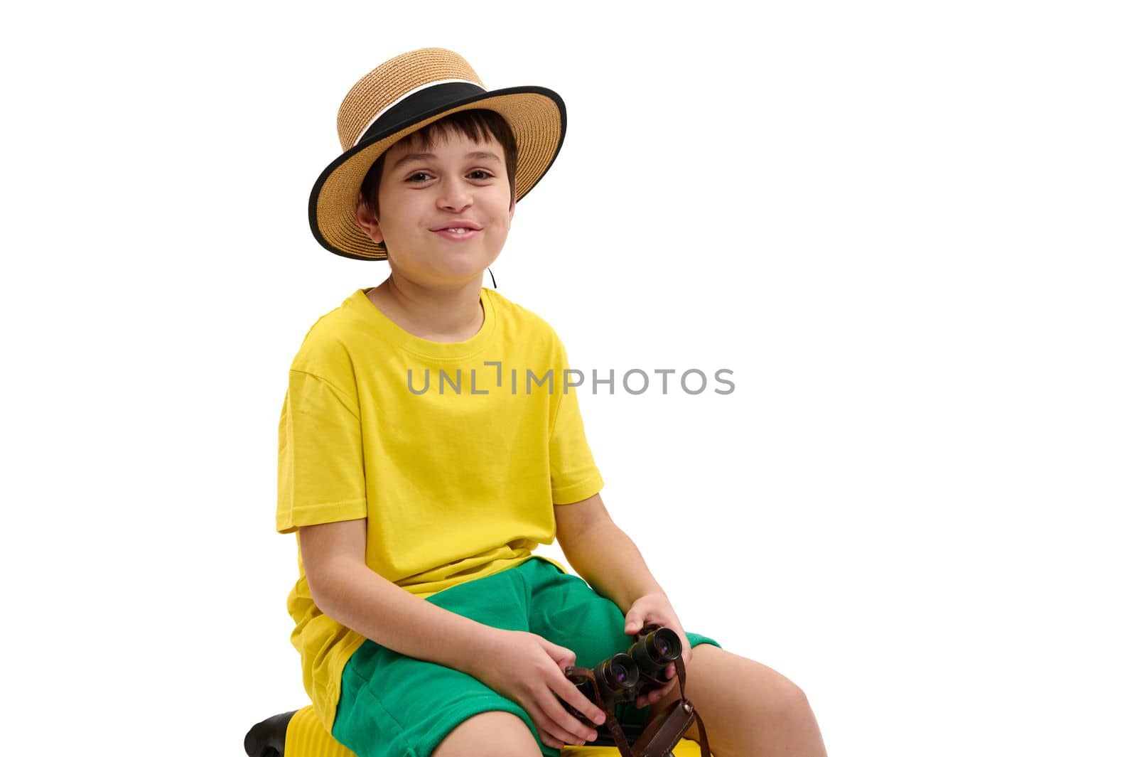 Smiling cheery traveler boy, wearing straw hat and yellow green summer wear, holding binoculars and smiling at camera, sitting on suitcase, isolated over white background. Copy advertising space