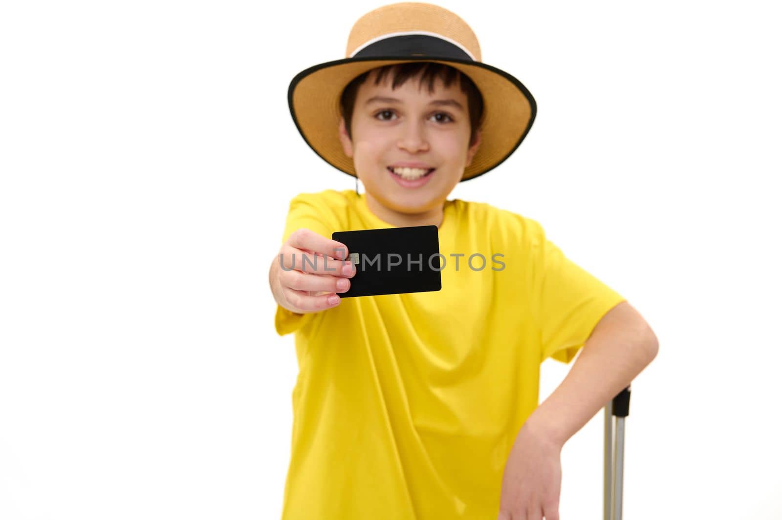 Close-up. Selective focus on the hand of a smiling child boy in summer straw hat and bright yellow t-shirt, holding out at camera a blank credit bank card with copy advertising space, white background