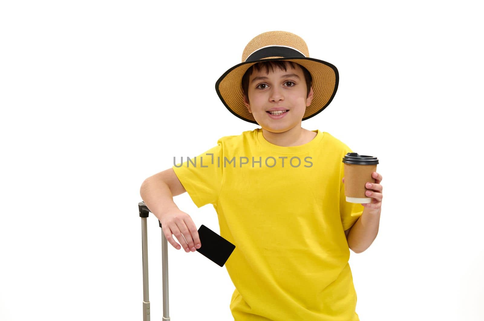 Adorable teenage boy - young tourist in bright yellow t-shirt and straw hat, standing over white background with blank plastic credit card, takeaway drink in paper cup and suitcase. Travel. Journey