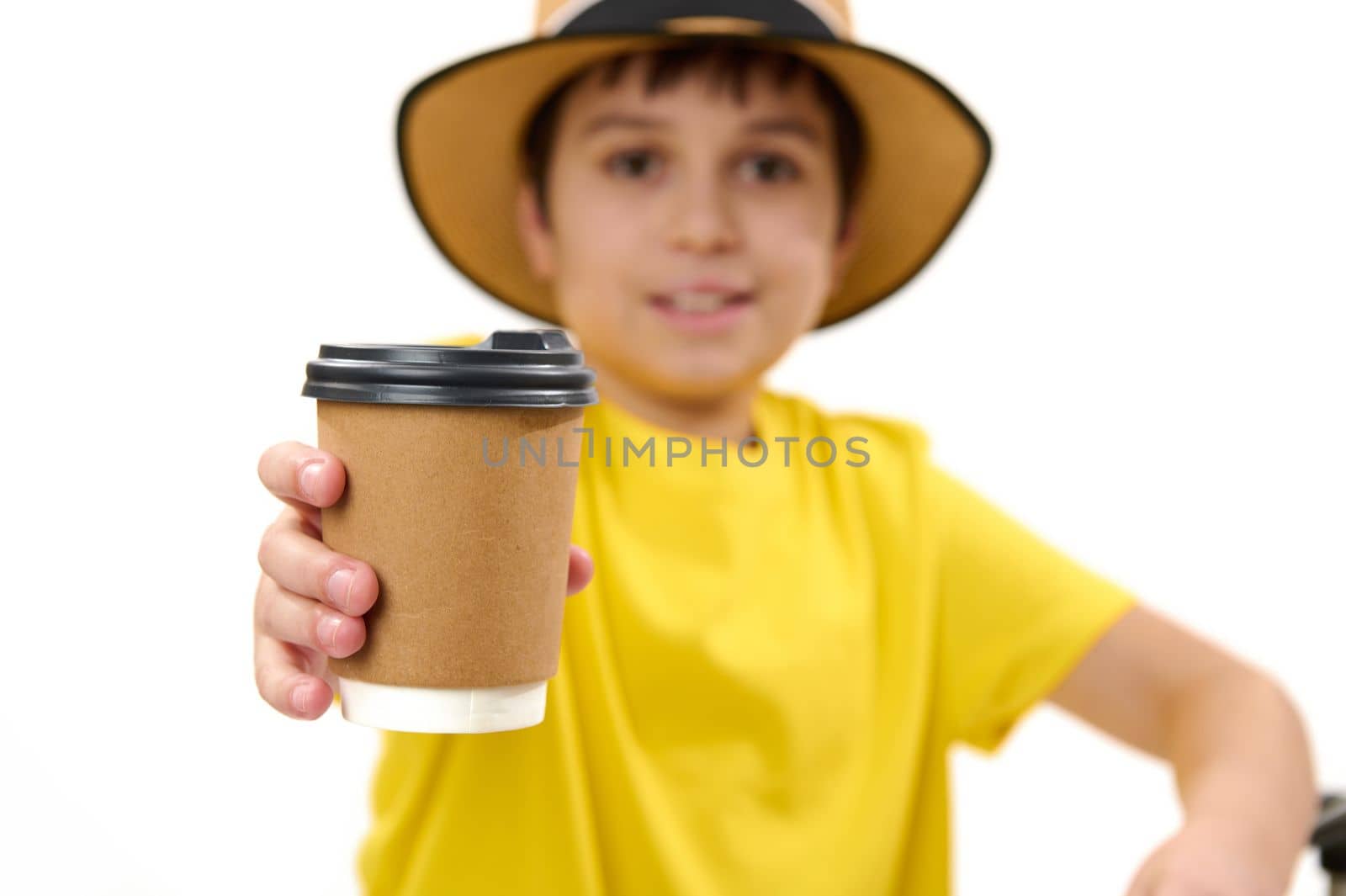 Selective focus on a disposable cardboard cup with takeaway hot drink in the hands of a blurred teenage boy in yellow t-shirt and straw hat, handing paper mug on camera, isolated over white background