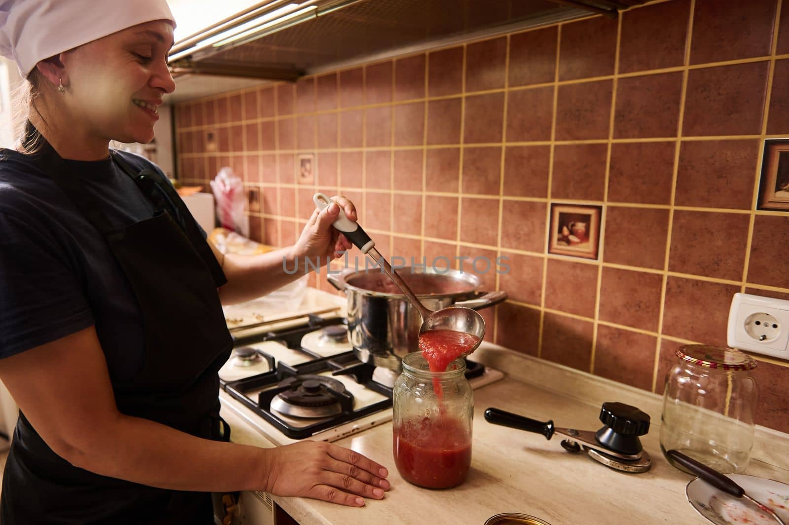 African American woman, pleasant housewife pouring freshly boiled tomato juice into a sterilized jar, preparing homemade organic passata or sauce. Preserving, canning or marinating food for the winter
