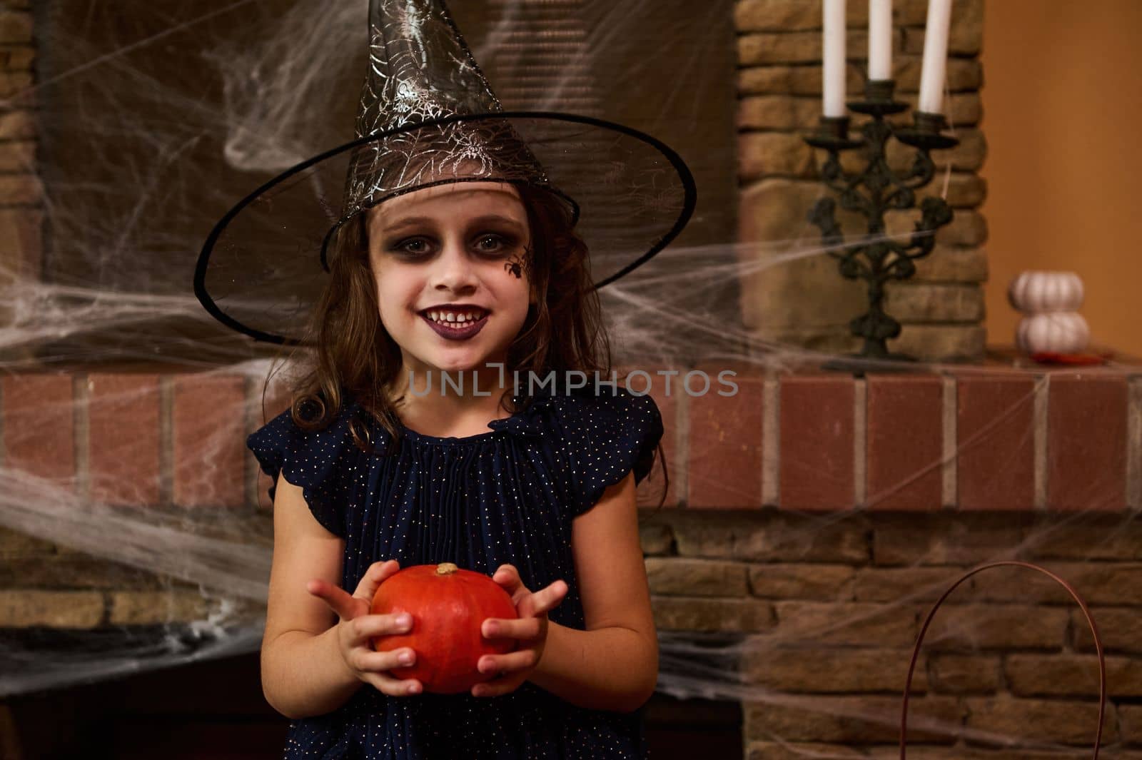 Little girl in witch dress and wizard hat with art make-up on her face, holding a pumpkin, smiling a toothy smile looking at the camera, sitting against a fireplace, decorated with spider web, candles
