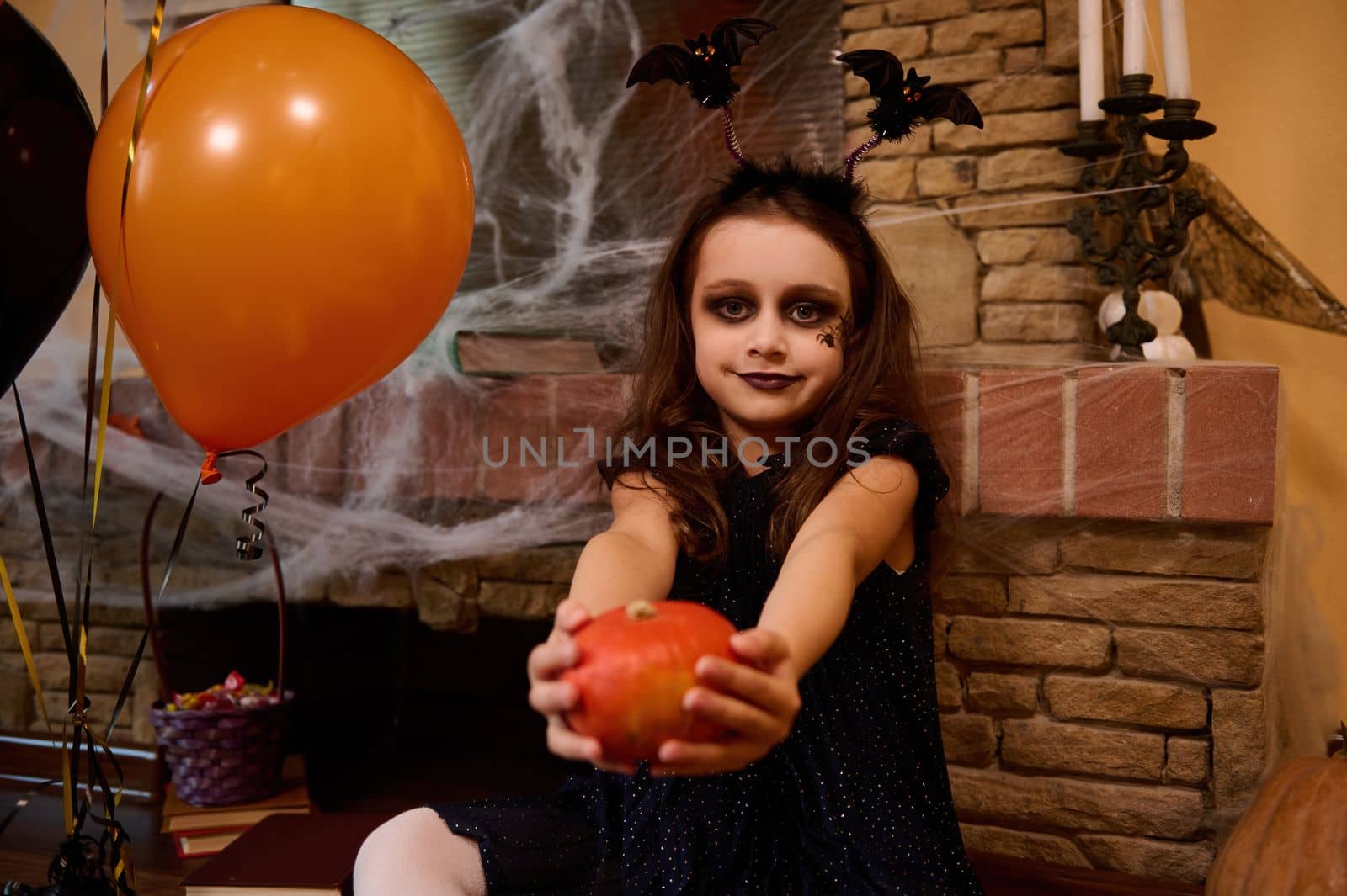 Caucasian gothic girl enchantress with black spider paint on her cheek and bat hoop, holds out a bright orange pumpkin, sits by a cobweb-covered fireplace and looks at the camera. Halloween home decor