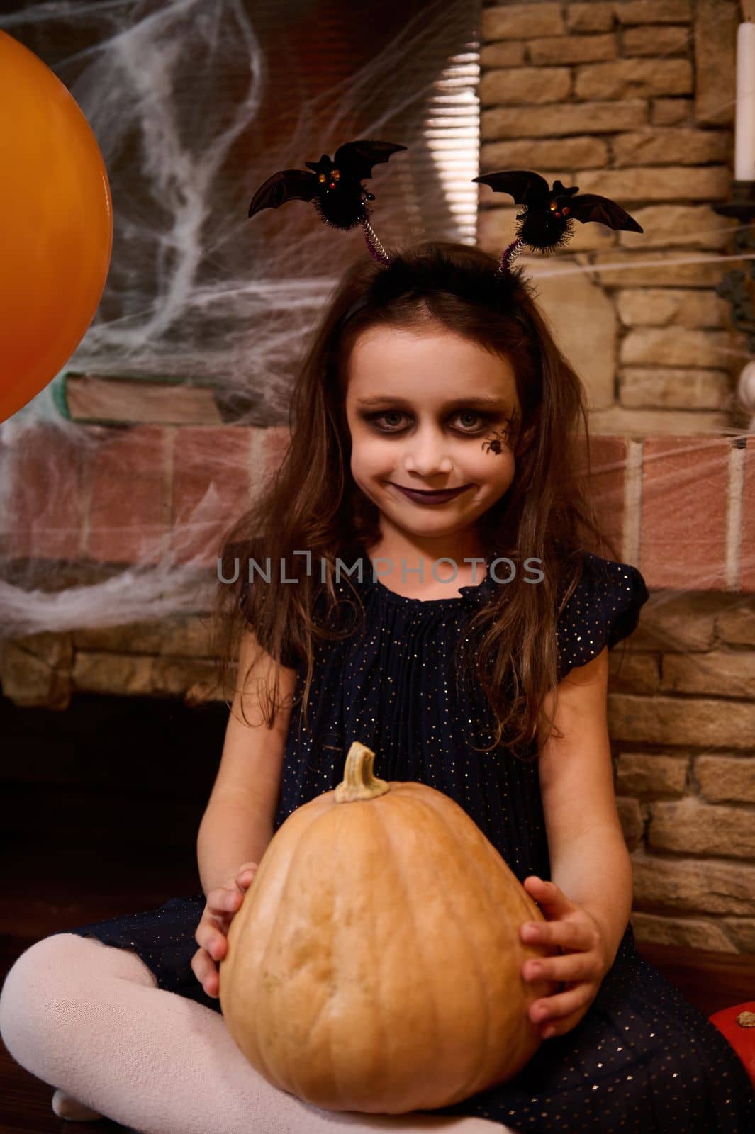 Little girl in black dress and a smokey eyes makeup, smiles looking at camera, making Halloween decoration at home by artgf