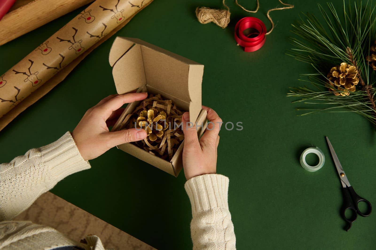 Top view of woman's hands packing a golden pine cone into a craft cardboard box over green background, surrounded by decorative wrapping materials and ornaments. Christmas and New Year preparations