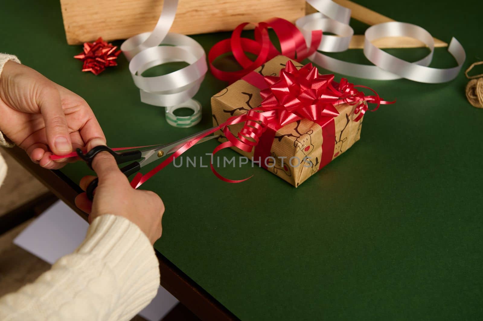 Close-up of woman's hands using scissors, cutting a red shiny ribbon in strips for making beautiful ornaments while wrapping presents for Christmas, over a green background with copy ad space