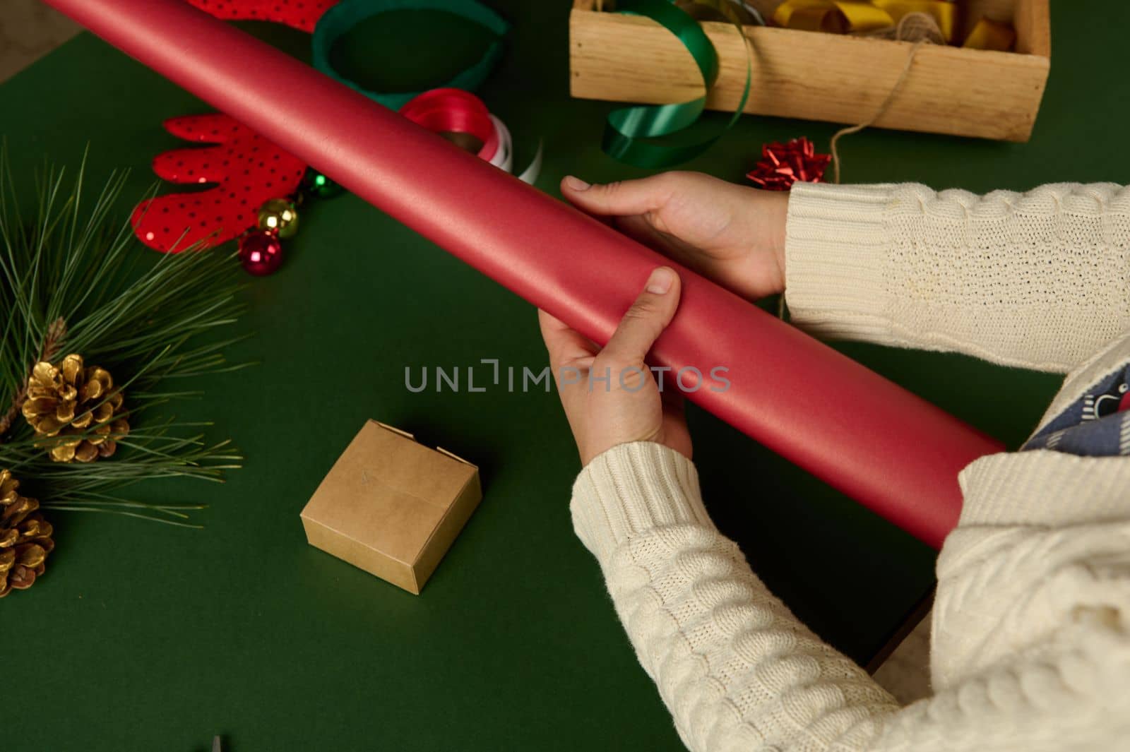 Top view woman's hands hold gift wrapping paper of red color while packing Christmas or New Year's presents, on green background. Festive decorations in wooden crate.Boxing Day. Xmas. December 25th