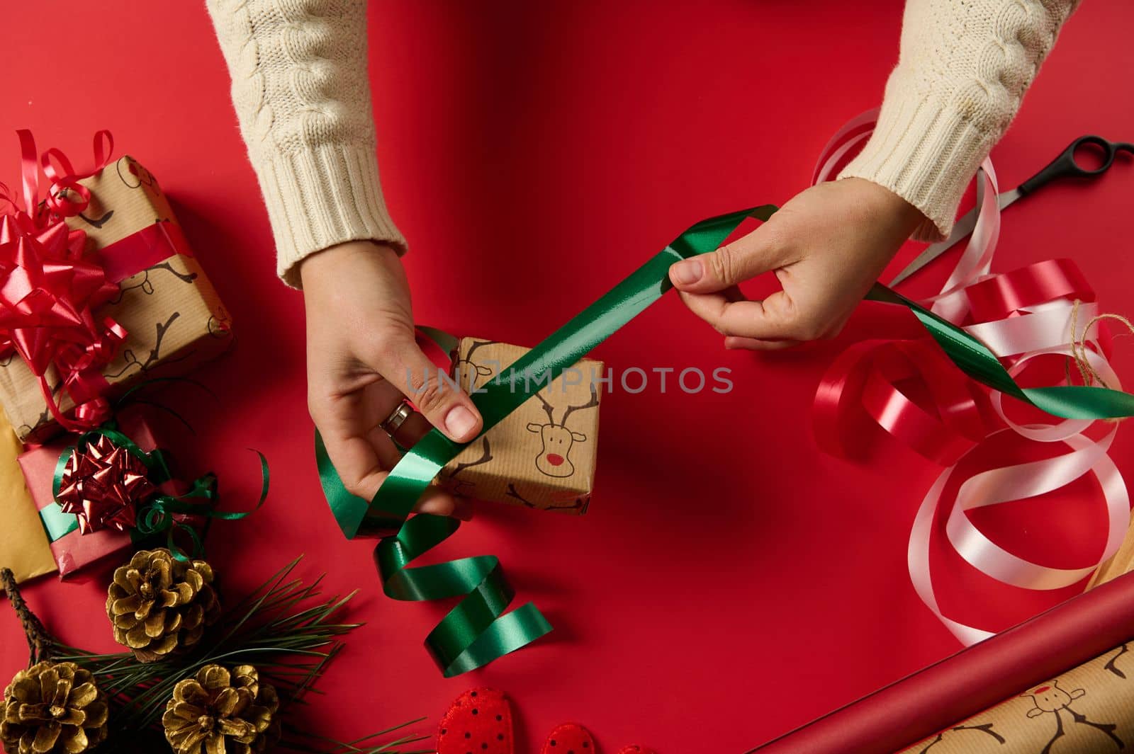 Christmas wrapping idea, xmas wrapping workspace. Female hands wrap gift box in paper and tying bow on red surface. Close Up woman wraps a present, decorates with shiny green ribbon and red tied bow