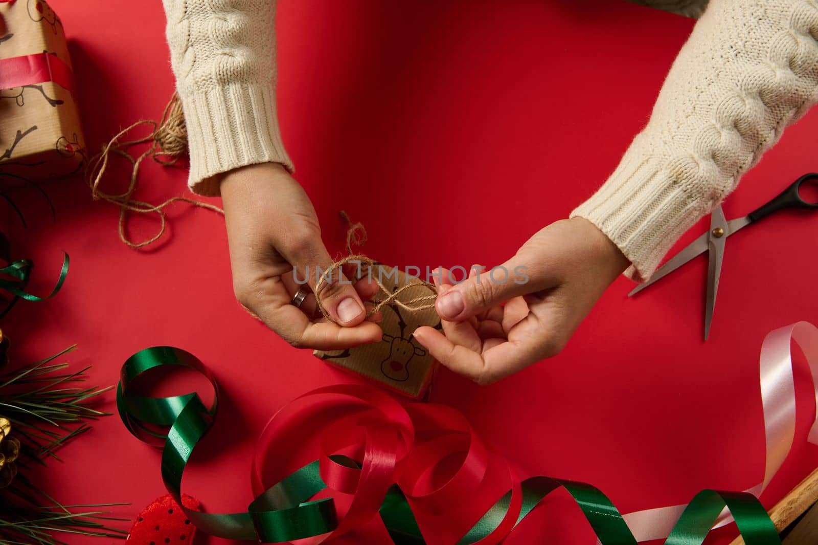 Top view craftswoman wrapping Christmas present in a decorative gift paper with deer pattern, tying a bow with craft linen rope. Boxing Day. Packing. Tying up gifts. Xmas and New Year preparations