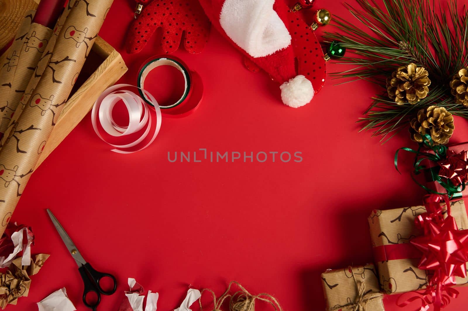 Top view of gift wrapping materials, shiny decorative ribbons and bows, scissors, boxes, Santa's hat lying in circle on red background with a copy advertising space in the center of the red background