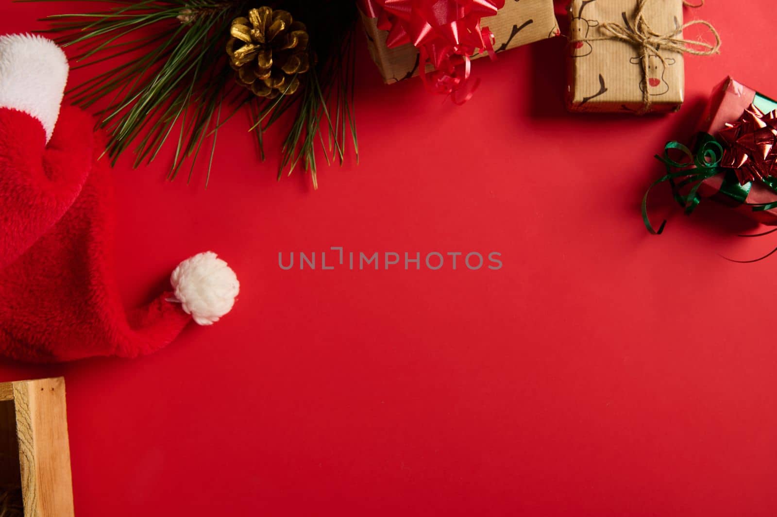 Christmas still life. View from above, partial view of Santa's hat, fir-tree branch, golden pine cones and Xmas gifts in the top of a bright red background with copy advertising space for text. Mockup