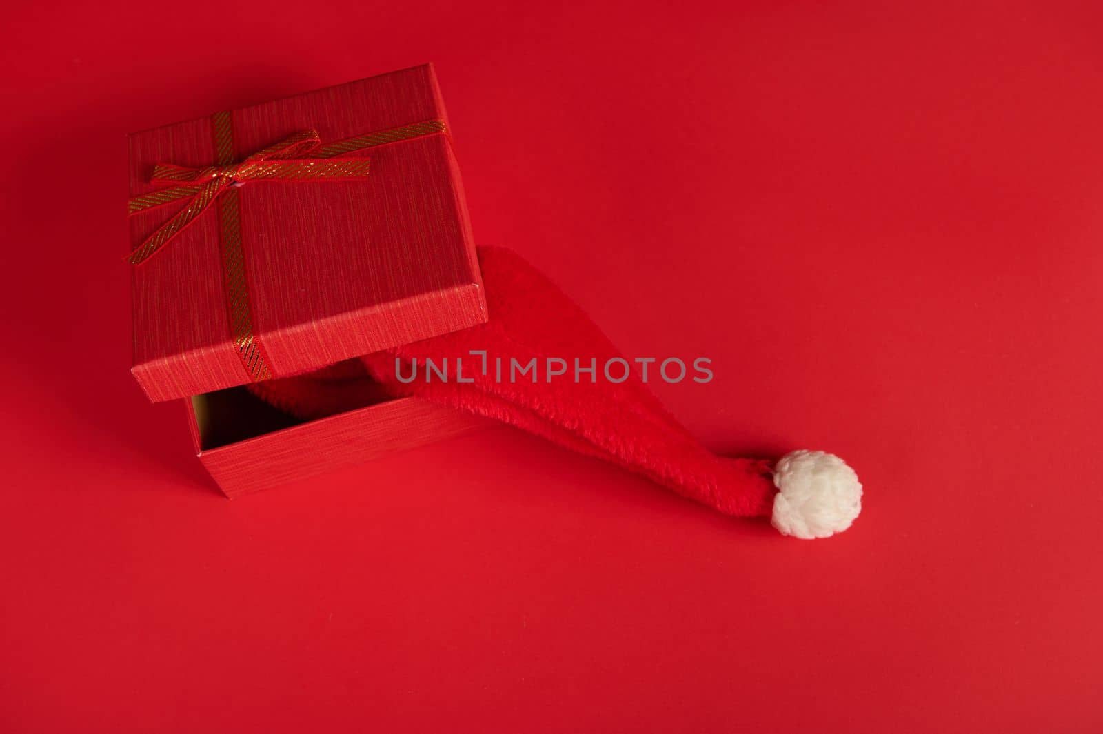 Top view of half open stylish red gift box, tied up with a red bow, with a Santa hat inside it, isolated over bright red background with copy ad space. New Year and Christmas preparations. Boxing Day