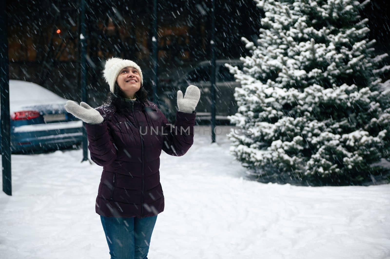 Charming cheerful multi-ethnic woman wearing wool mittens and warm clothes, looking up the evening sky, enjoys the snowfall in the city
