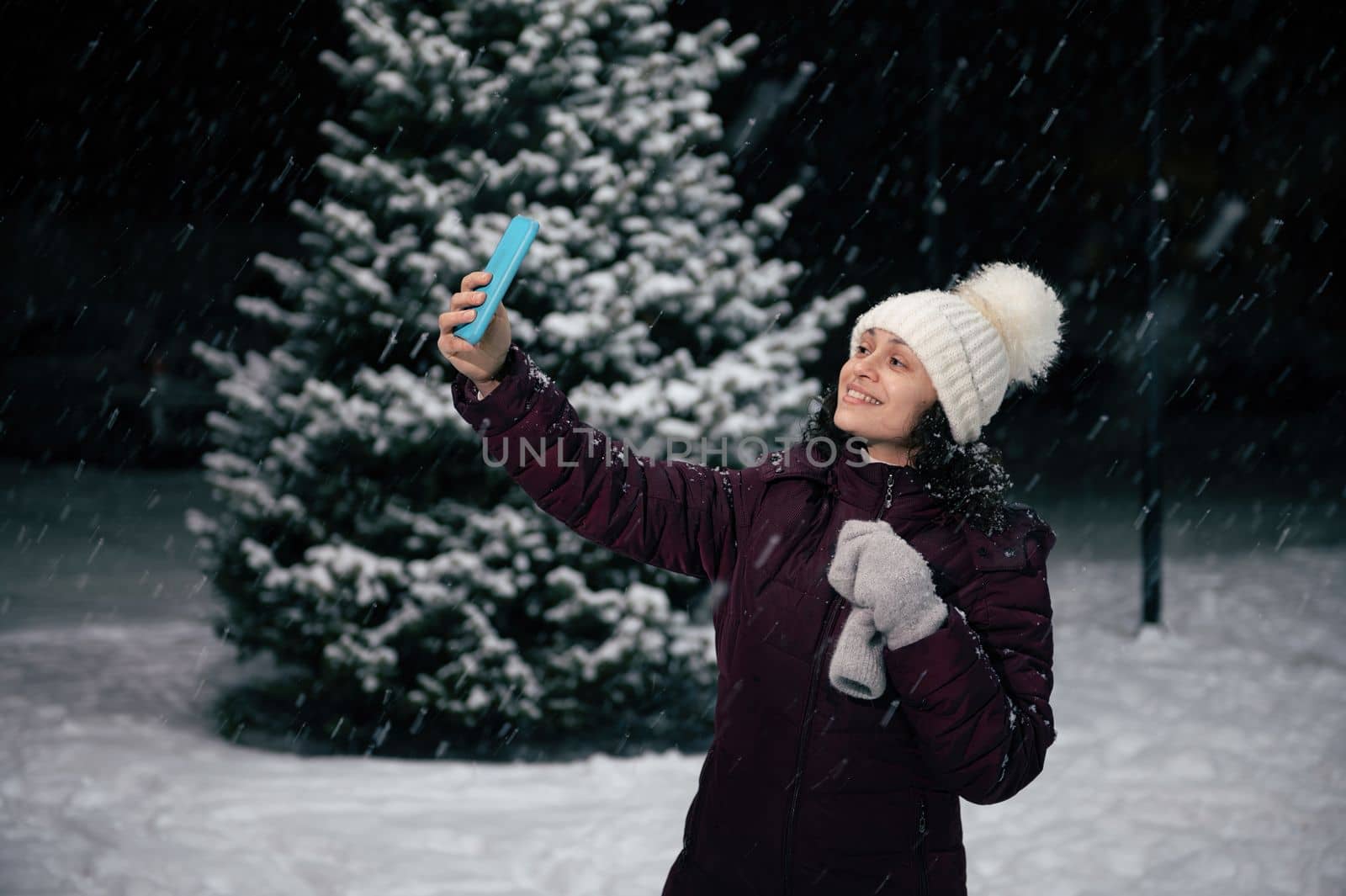 Multi-ethnic pretty woman in warm clothes, white wool mittens and hat, smiles a beautiful toothy smile while taking selfie on her smartphone, enjoying a snowy winter evening. Merry Christmas concept