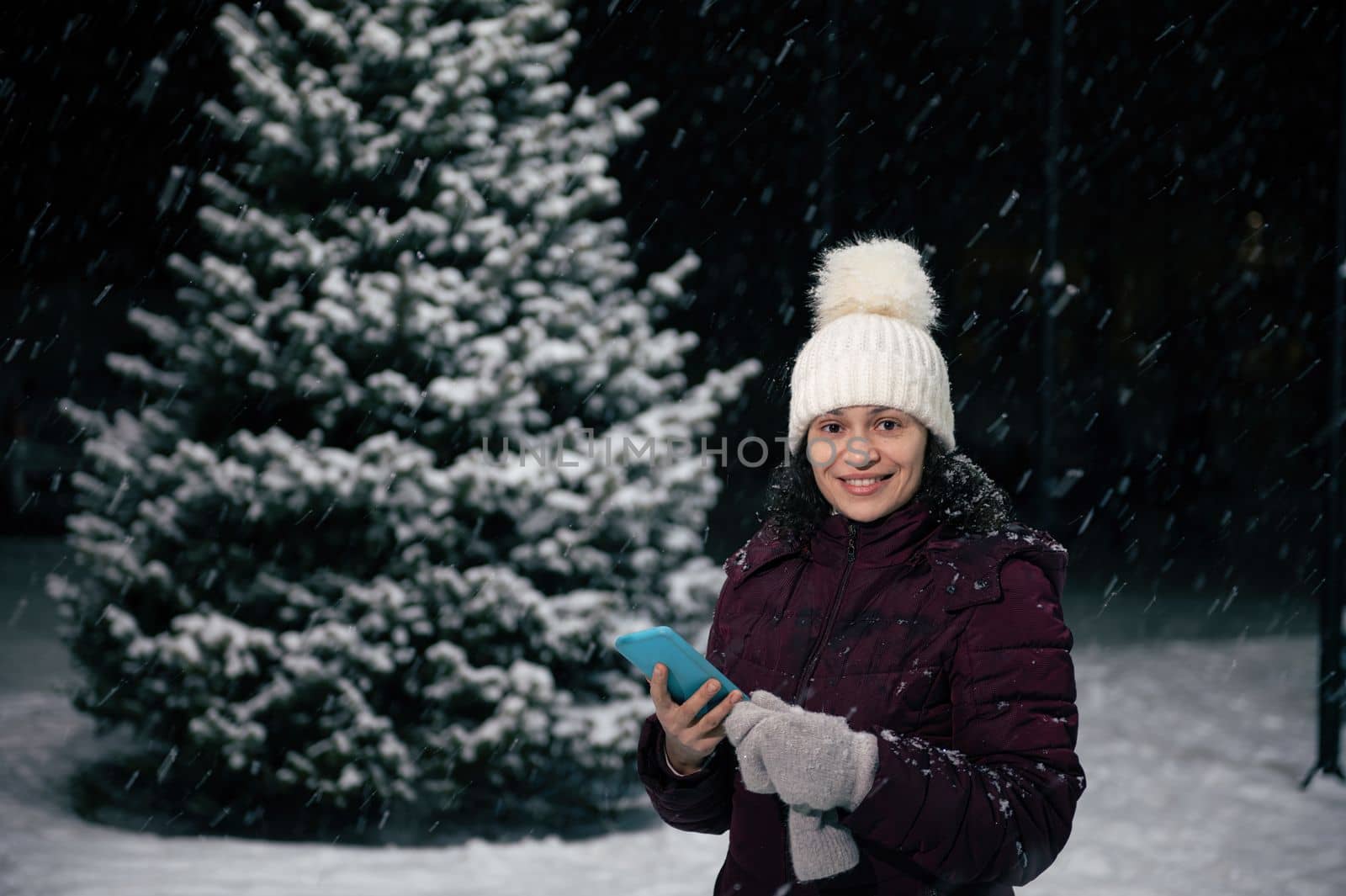 Beautiful Latin American woman in warm clothes, wool mittens and hat, holding smartphone and smiling a toothy smile looking at camera, standing against a tall fir-tree in the snow covered city street