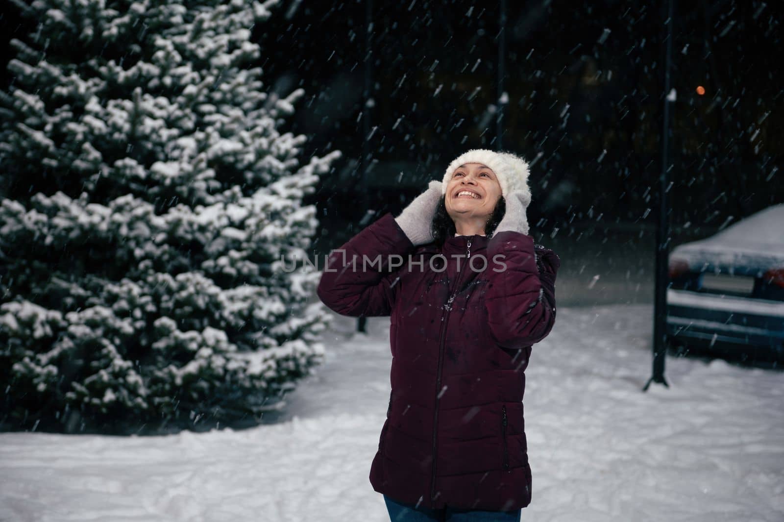 Happy cheerful multi-ethnic woman in warm clothes, having fun outdoor, smiling a toothy smile looking up the sky, enjoys a snowfall on a snow covered city. Winter holidays. Merry Christmas concept