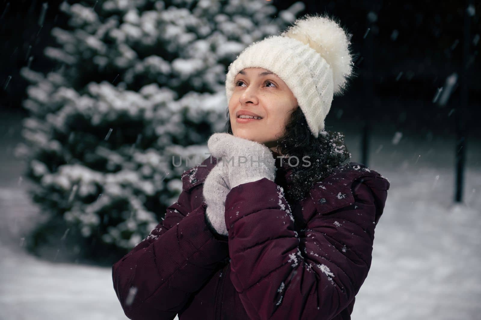 Close-up portrait of charming dark-haired multi-ethnic woman in warm clothes, white wool hat and mittens in the snow covered nature. Snowfall. Winter holidays. Happy New Year. Mmerry Christmas concept