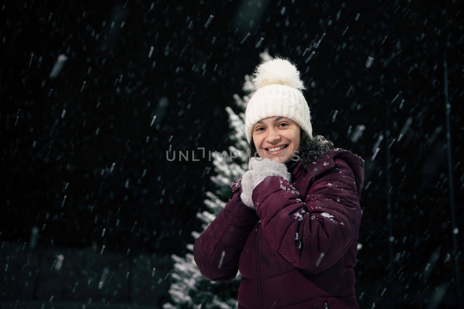 Portrait of a charming dark-haired multi-ethnic woman in warm clothes, white wool hat and mittens, smiling a beautiful toothy smile looking at the camera, standing in the snow covered nature.