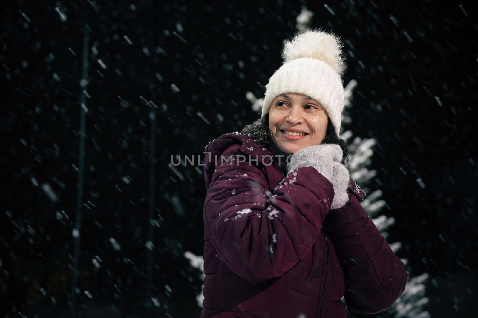 Portrait of a smiling beautiful multi-ethnic woman in a warm wool hat and mittens on a city street on a snowy evening. Snowfall. Merry Christmas and winter holidays concept