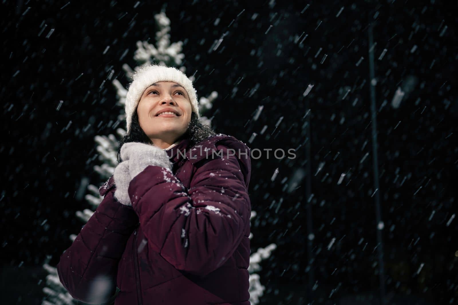 Portrait of delightful beautiful woman in warm clothes, looking up to the sky, enjoying christmas atmosphere outdoors on snowy evening. Snowfall. Winter holidays and Merry Christmas concept