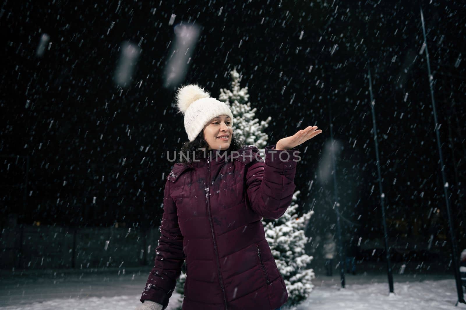 Charming happy woman in warm clothes, catching snowflakes on her hand while walking down the city street, on a snowy evening. Winter. Snowfall. Merry Christmas and Happy New Year concept