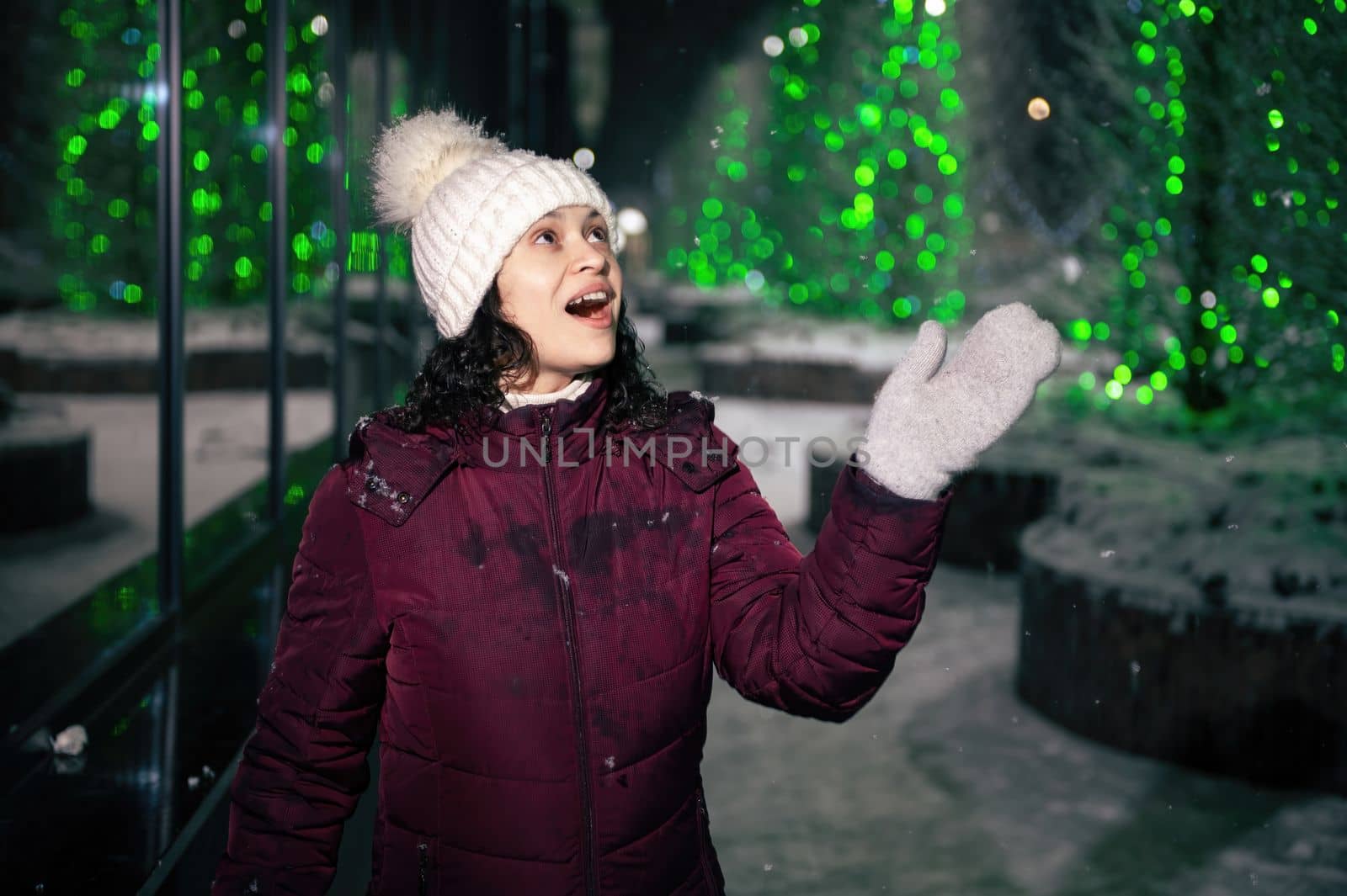 Pretty multi-ethnic woman in warm clothes, catches snowflakes, enjoys walking down the city street, lightened by garlands on a snowy winter night. Winter holidays. Snowfall. Merry Christmas