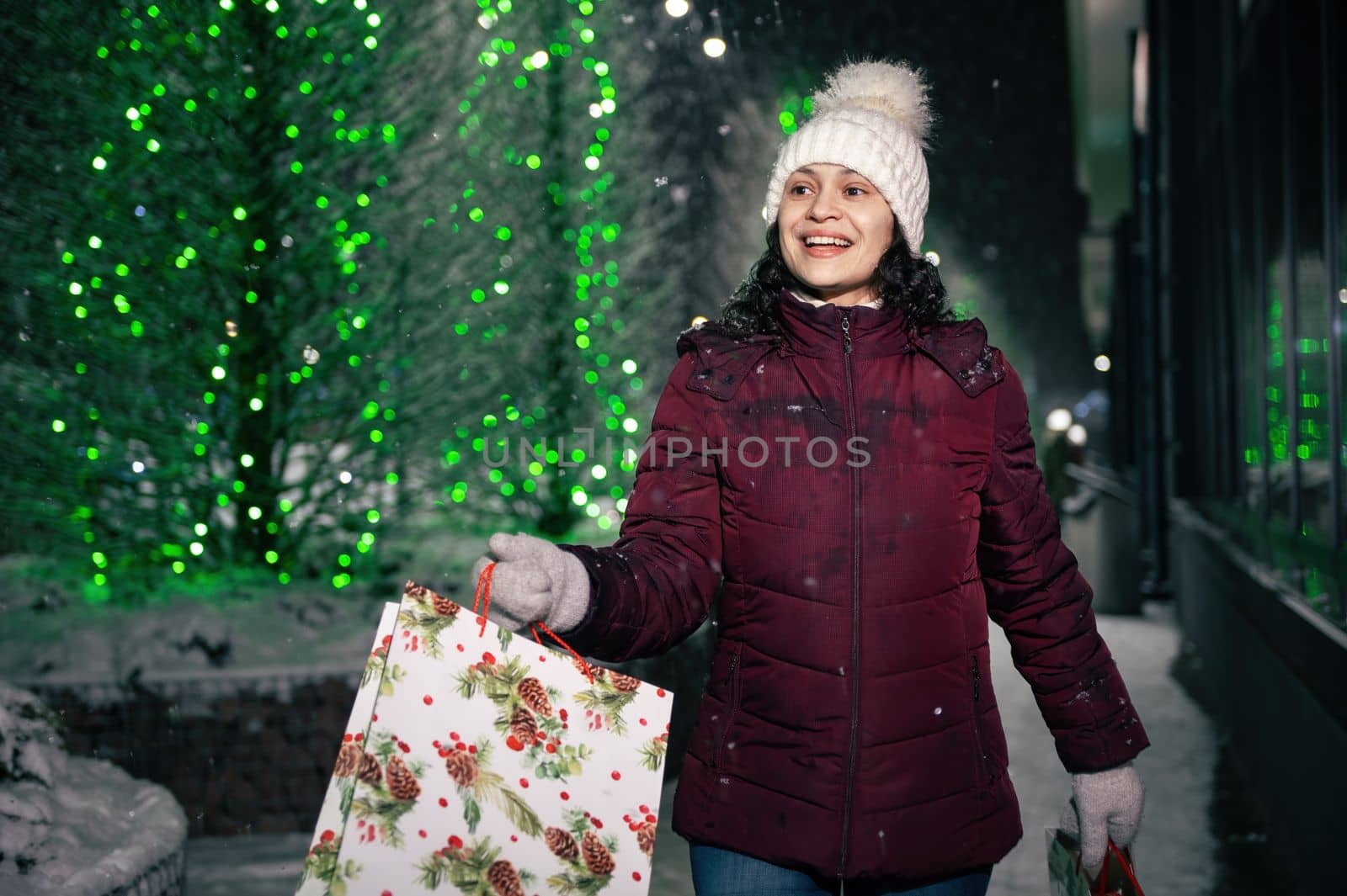 Beautiful multi-ethnic woman walking with shopping bags on a winter street, illuminated by garlands at snowy winter night. Merry Christmas and Happy winter holidays concept