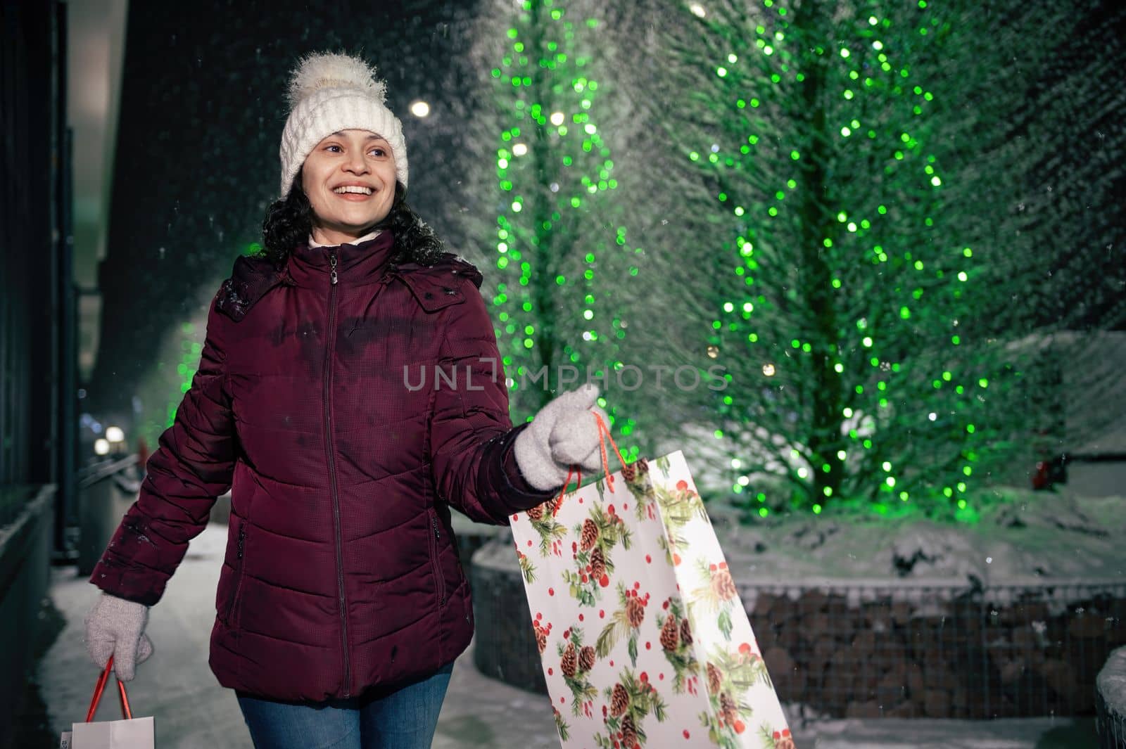 Delightful Latin American woman walking with shopping bags on a city street, illuminated by garlands at snowy winter night. Christmas and New Year concept
