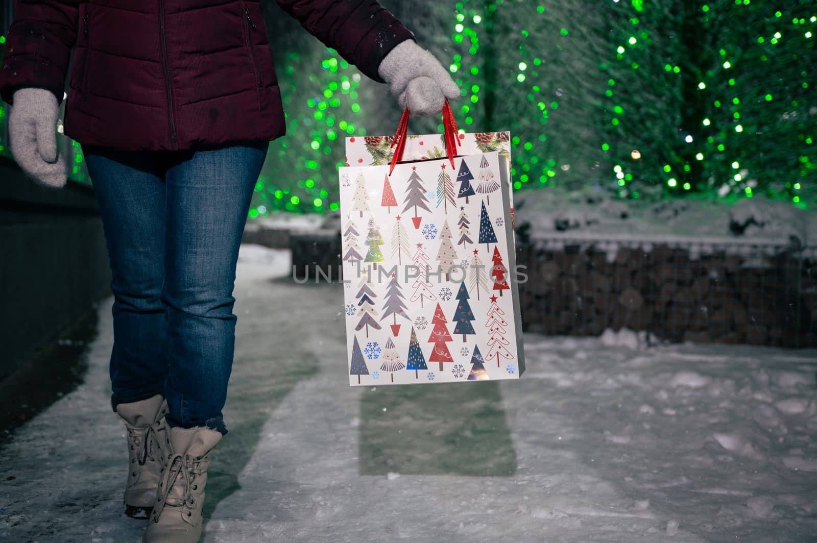 Selective focus on shopping bags with Christmas pattern in the hands of a pretty woman, walking down the snow covered city street, on a winter night, against fir-trees illuminated by festive garlands